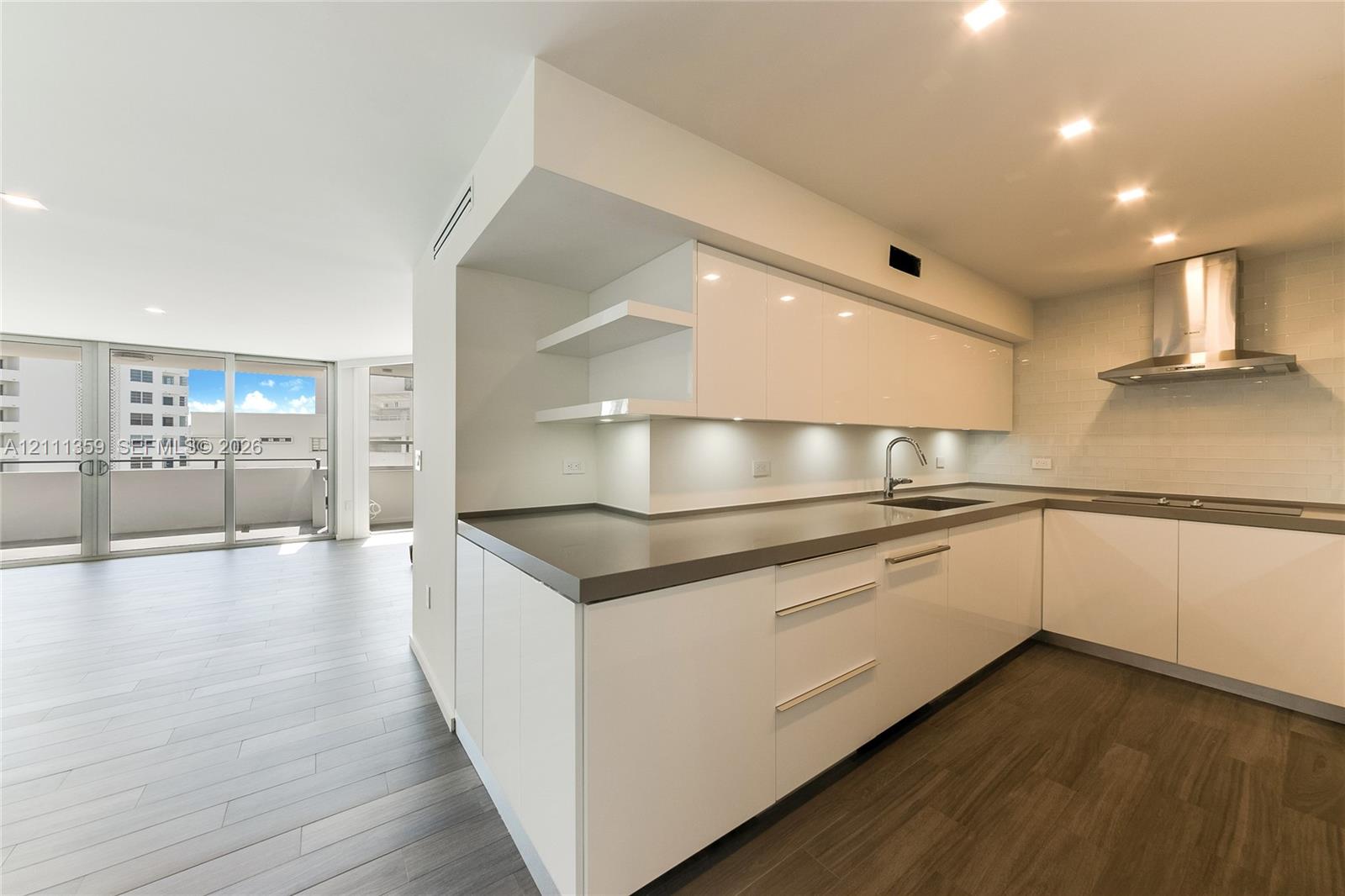 Modern kitchen featuring high-gloss white cabinetry, solid-surface countertops, stainless steel range hood, recessed lighting, and wood-finish flooring