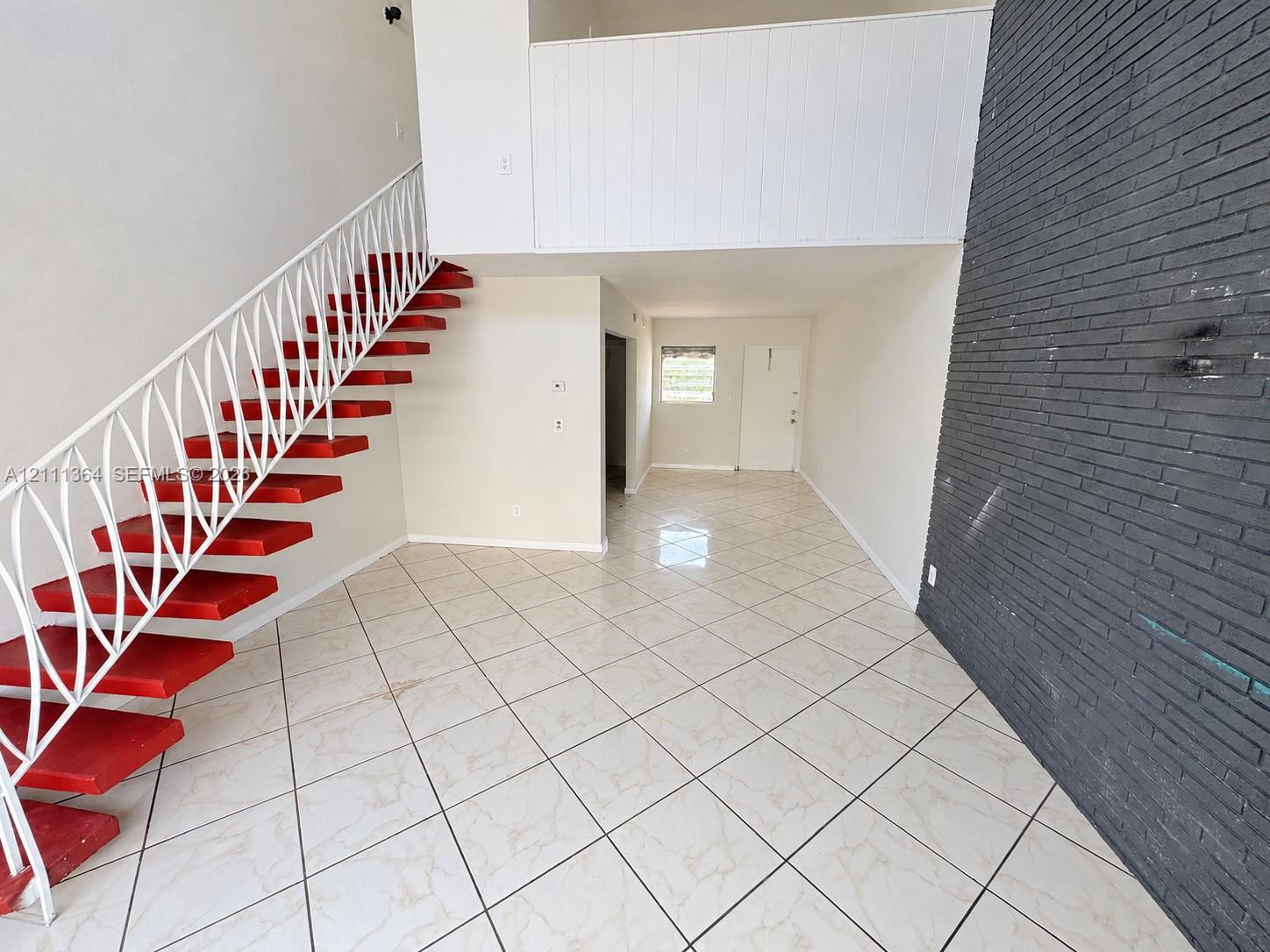 Open floor plan featuring high ceilings, a floor-to-ceiling dark brick accent wall, tile flooring in a diamond pattern, and a staircase with open risers and metal latticework railing