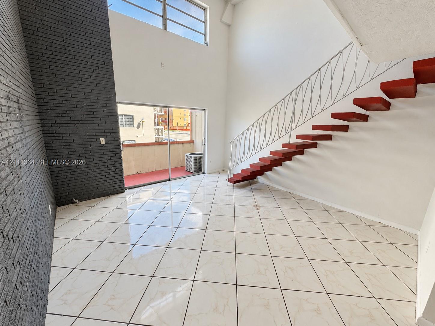 Double-height ceiling, decorative dark-toned brick accent wall, ceramic tile flooring, floating staircase with custom iron railing, and sliding glass doors Double-height ceiling, decorative dark-toned brick accent wall, ceramic tile flooring, floating staircase with custom iron railing, and sliding glass doors
