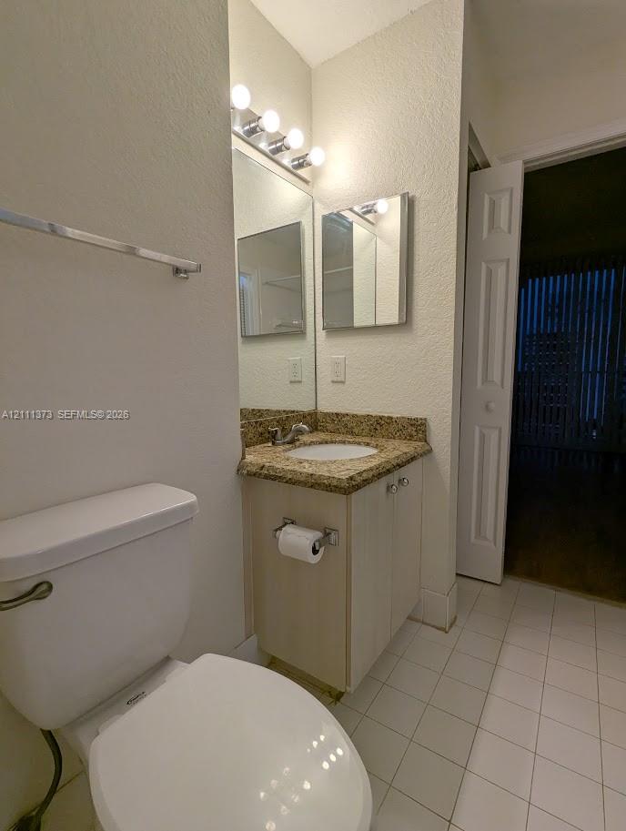 Single-sink vanity with a stone-finish countertop, white cabinetry, wall-mounted mirror, four-bulb vanity lighting, and white tile flooring