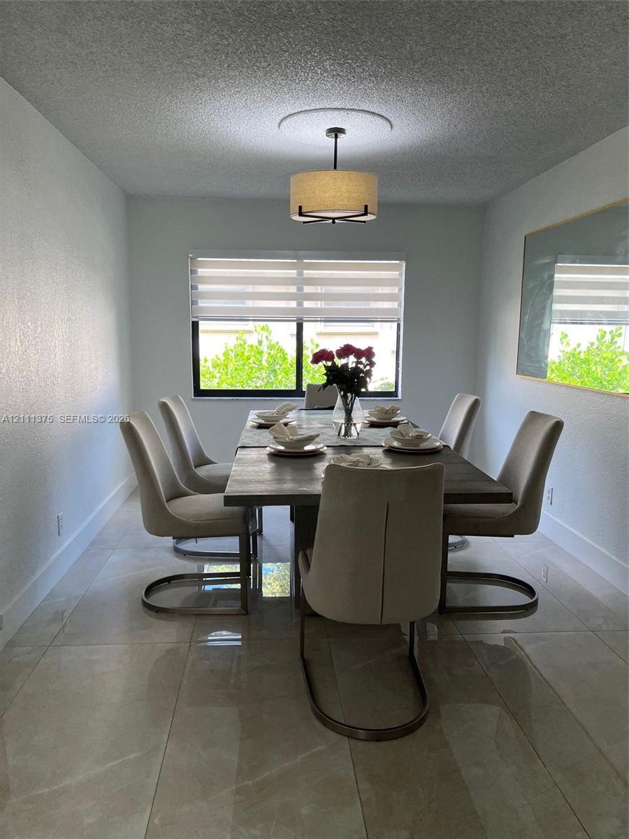 Dining area featuring large-format tile flooring, textured ceiling, drum-style pendant light with medallion, and window equipped with dual-layer fabric shades