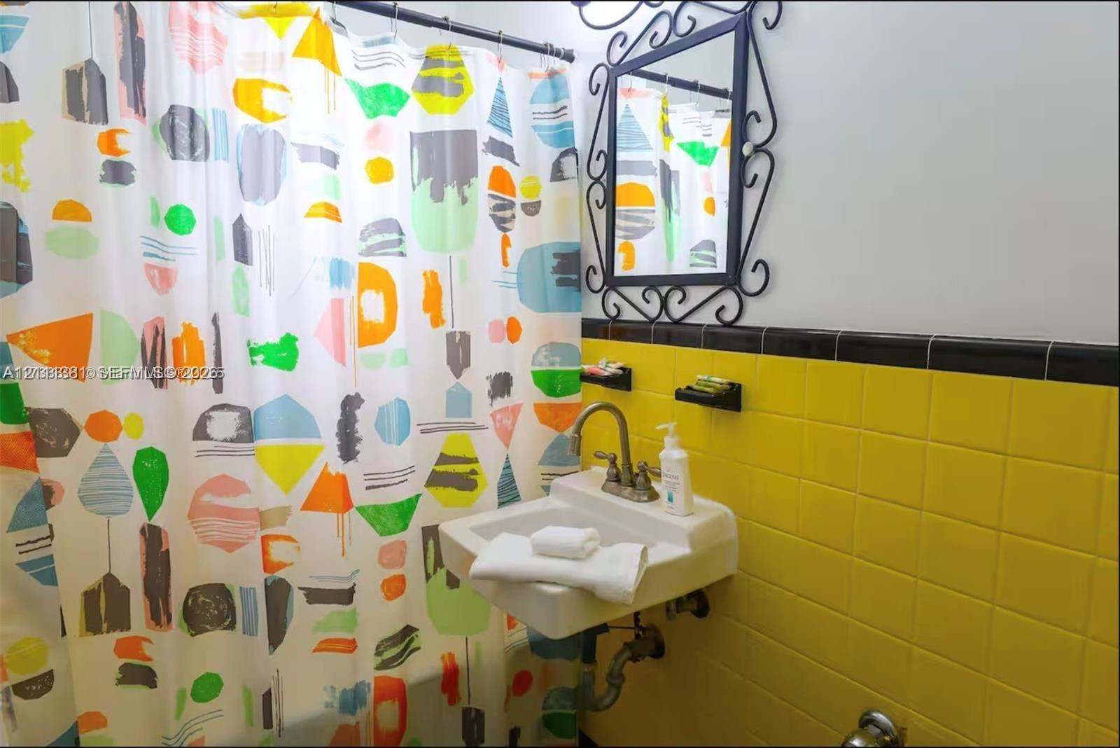 Wall-mounted porcelain sink with dual-handle metal faucet, yellow ceramic tile wainscoting with black trim, and decorative wrought-iron framed wall mirror