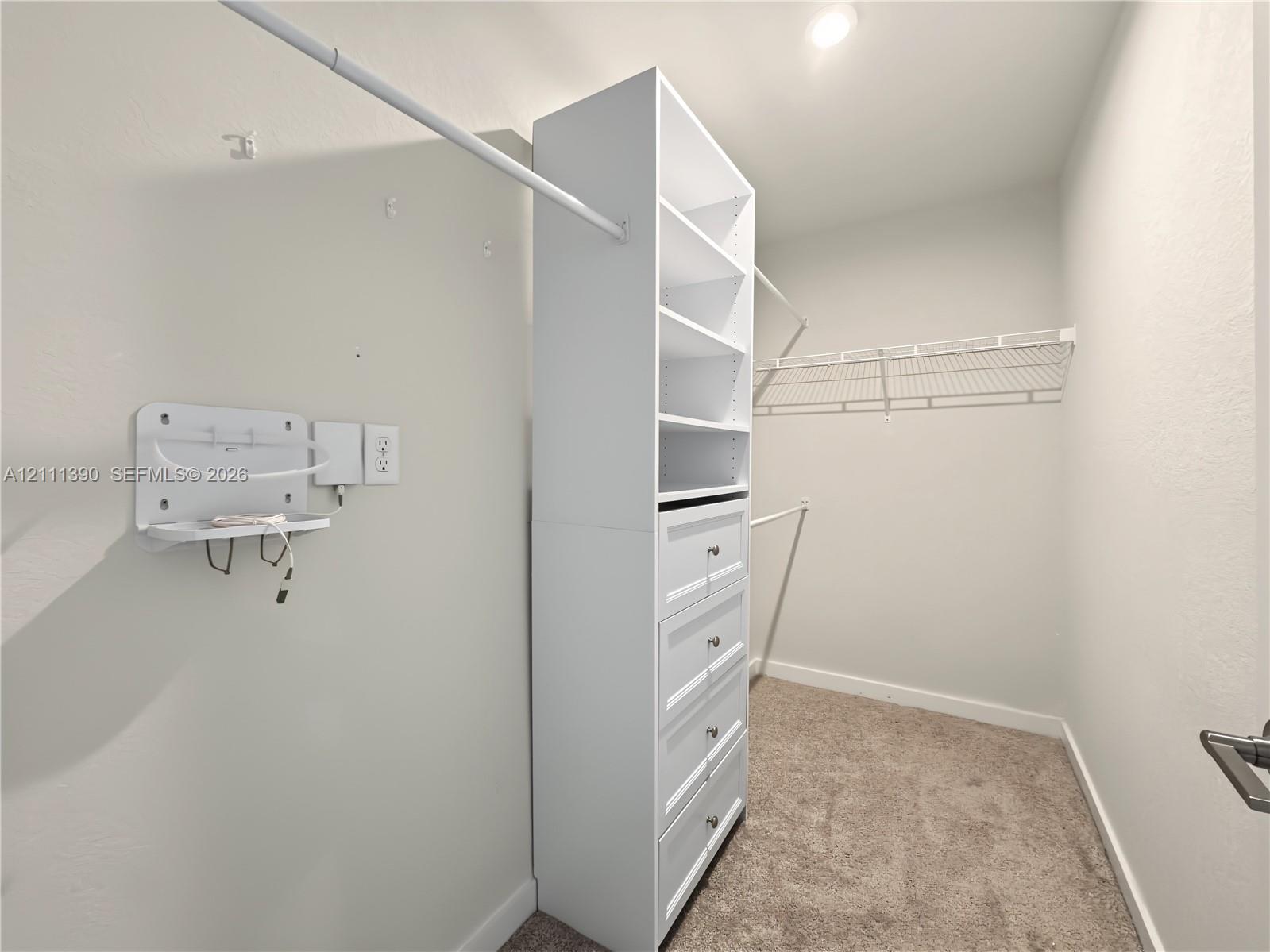 Walk-in closet featuring white custom built-in shelving, a five-drawer dresser unit, wall-mounted wire shelving, and neutral carpeting
