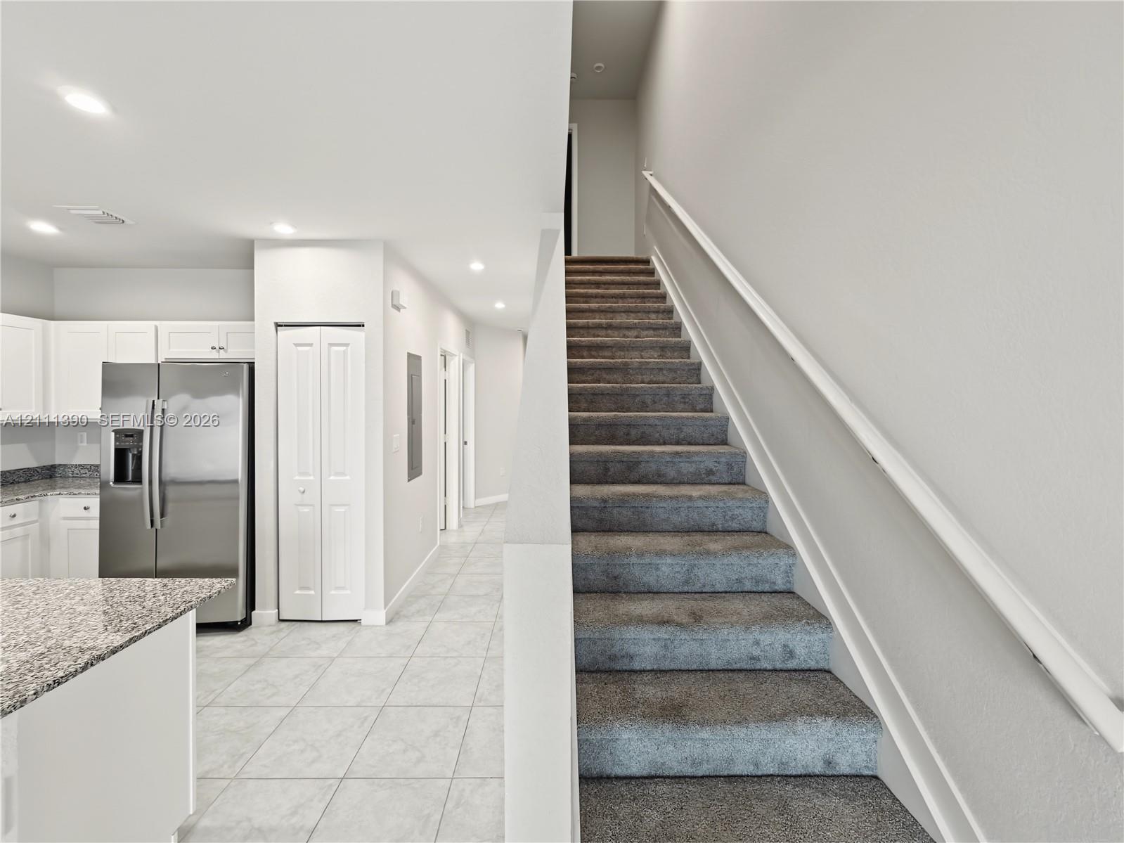 Kitchen featuring stainless steel refrigerator, white cabinetry, and stone-finish countertops, with tiled flooring and carpeted staircase with white handrail