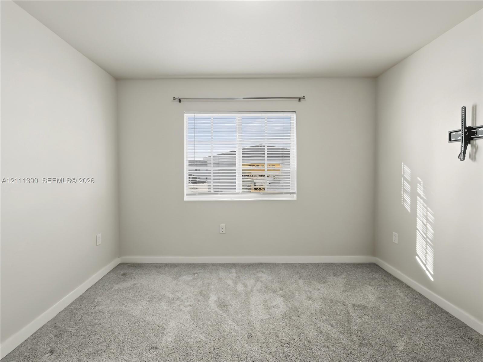 Neutral-toned interior room featuring wall-to-wall carpeting, a horizontal sliding window with attached curtain rod, and a wall-mounted television bracket