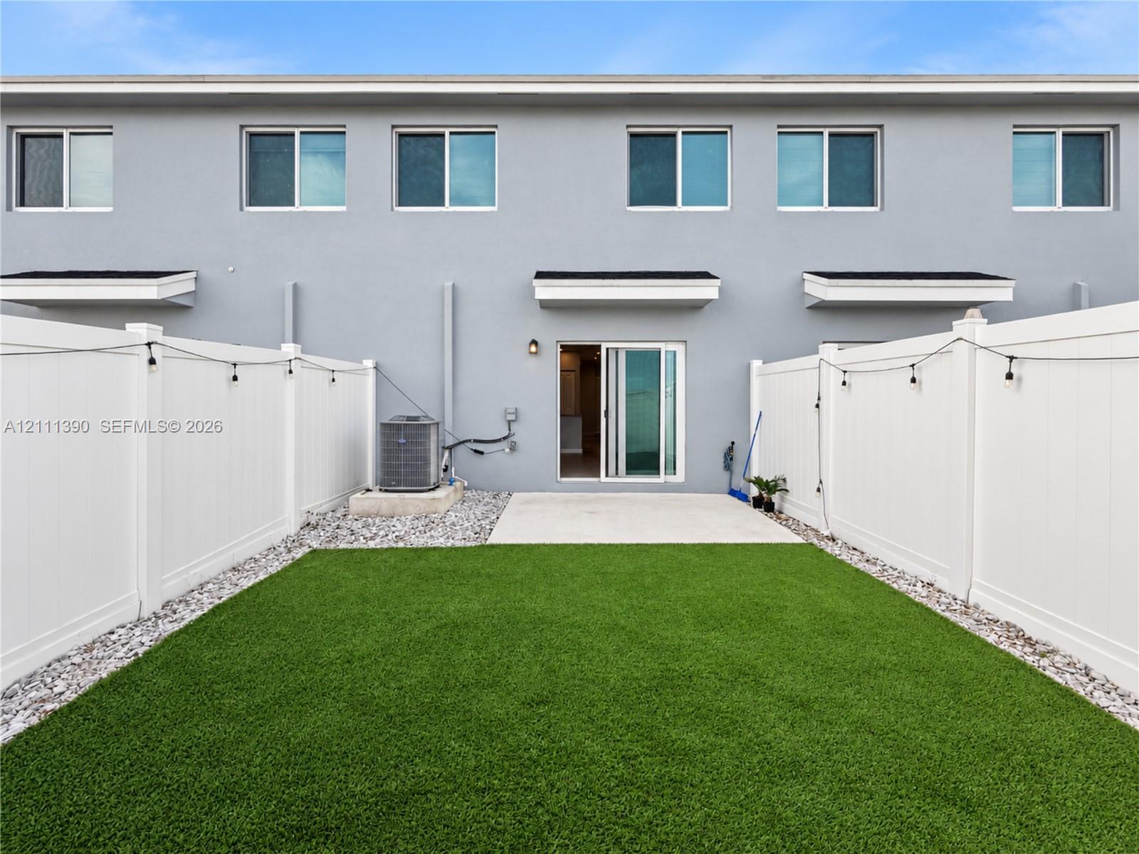 Private backyard featuring synthetic turf, concrete patio, white vinyl privacy fencing, and exterior sliding glass door