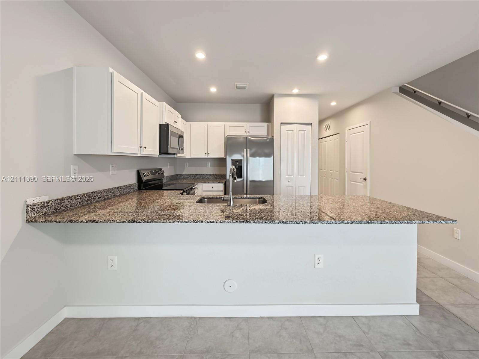 Kitchen featuring white shaker-style cabinetry, stone-finish countertops, stainless steel appliances, a peninsula with an undermount sink, and tile flooring