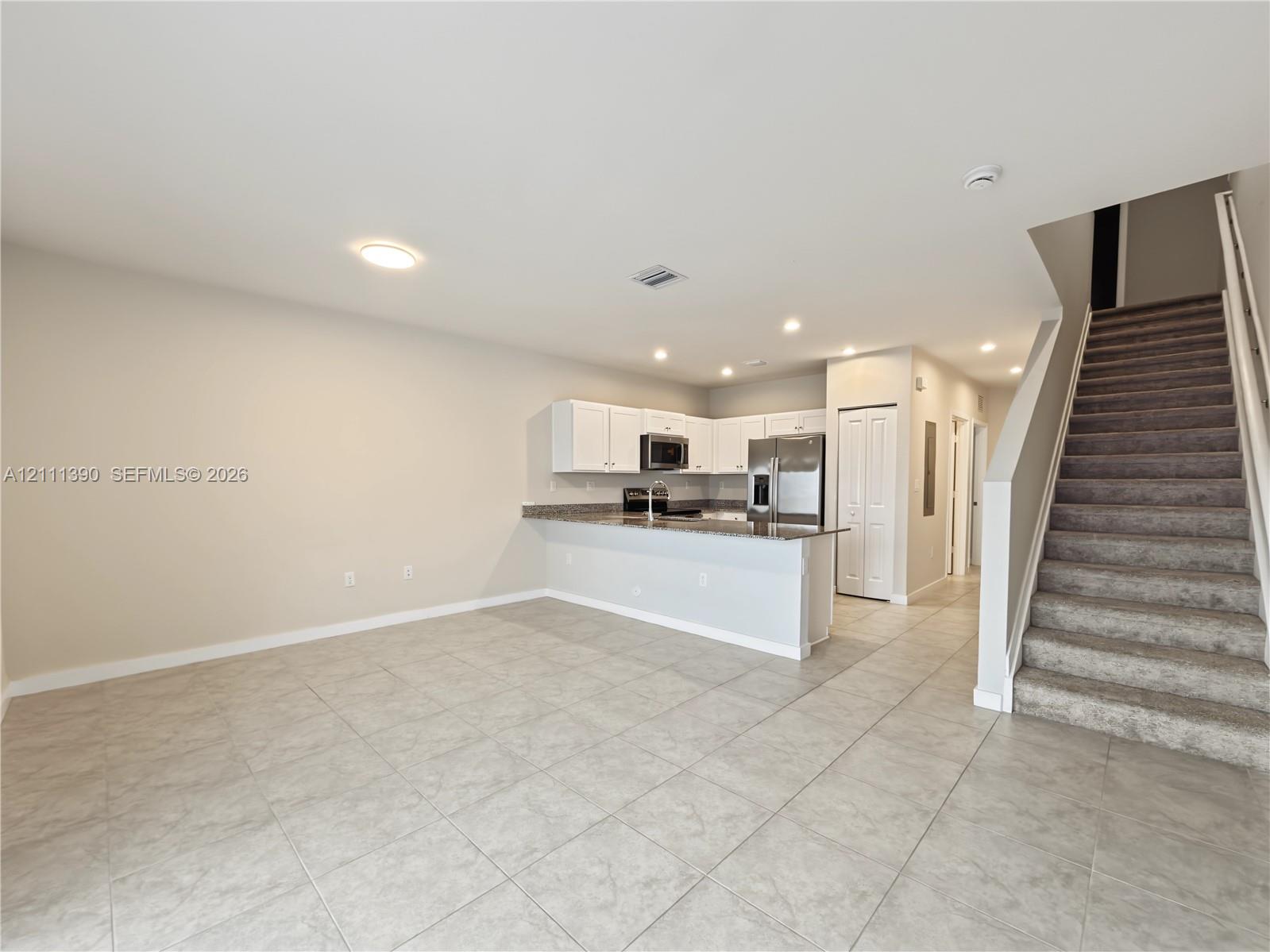 Open-concept kitchen featuring white cabinetry, stone-finish countertops, stainless steel appliances, and tile flooring, with carpeted stairs