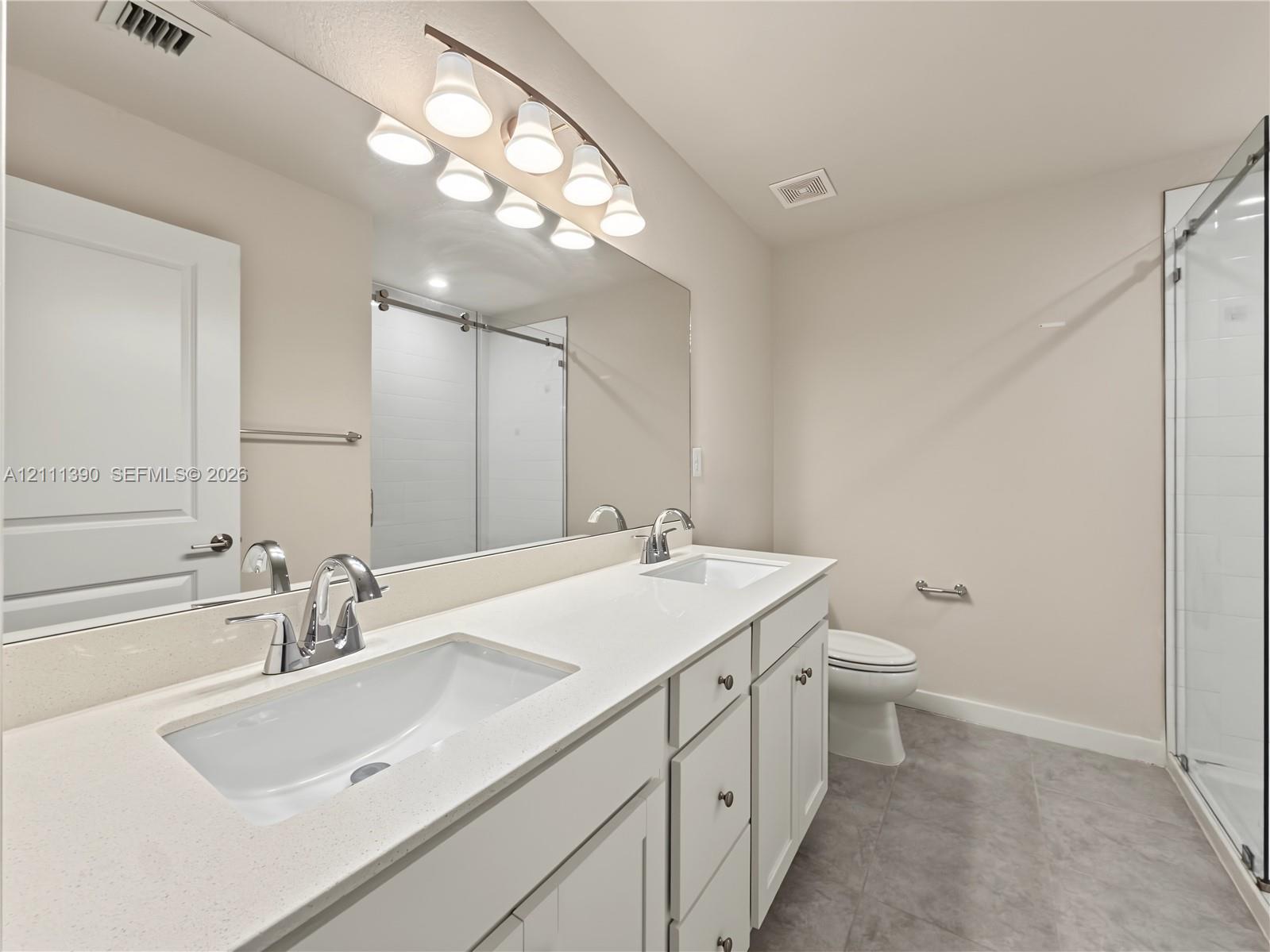 Dual-sink vanity with white cabinetry, light-toned stone countertops, polished chrome fixtures, full-width mirror, and a glass-enclosed shower