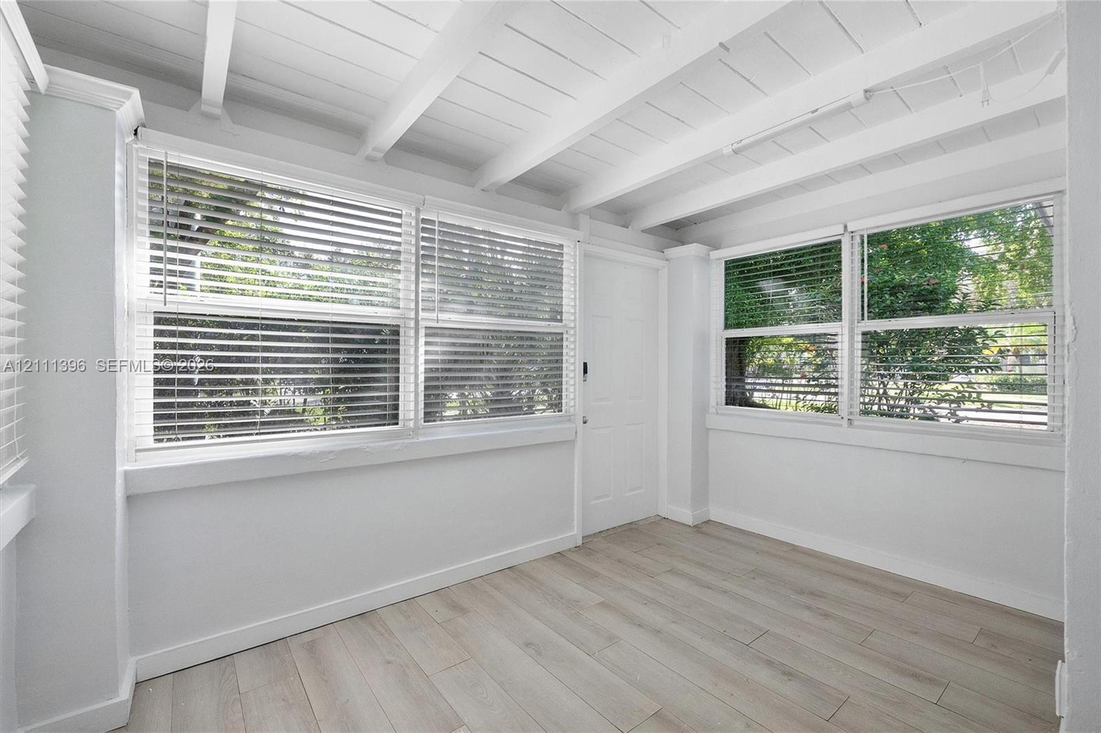Exposed beamed ceiling with white finish, wood-finish flooring, and multiple large windows with horizontal blinds