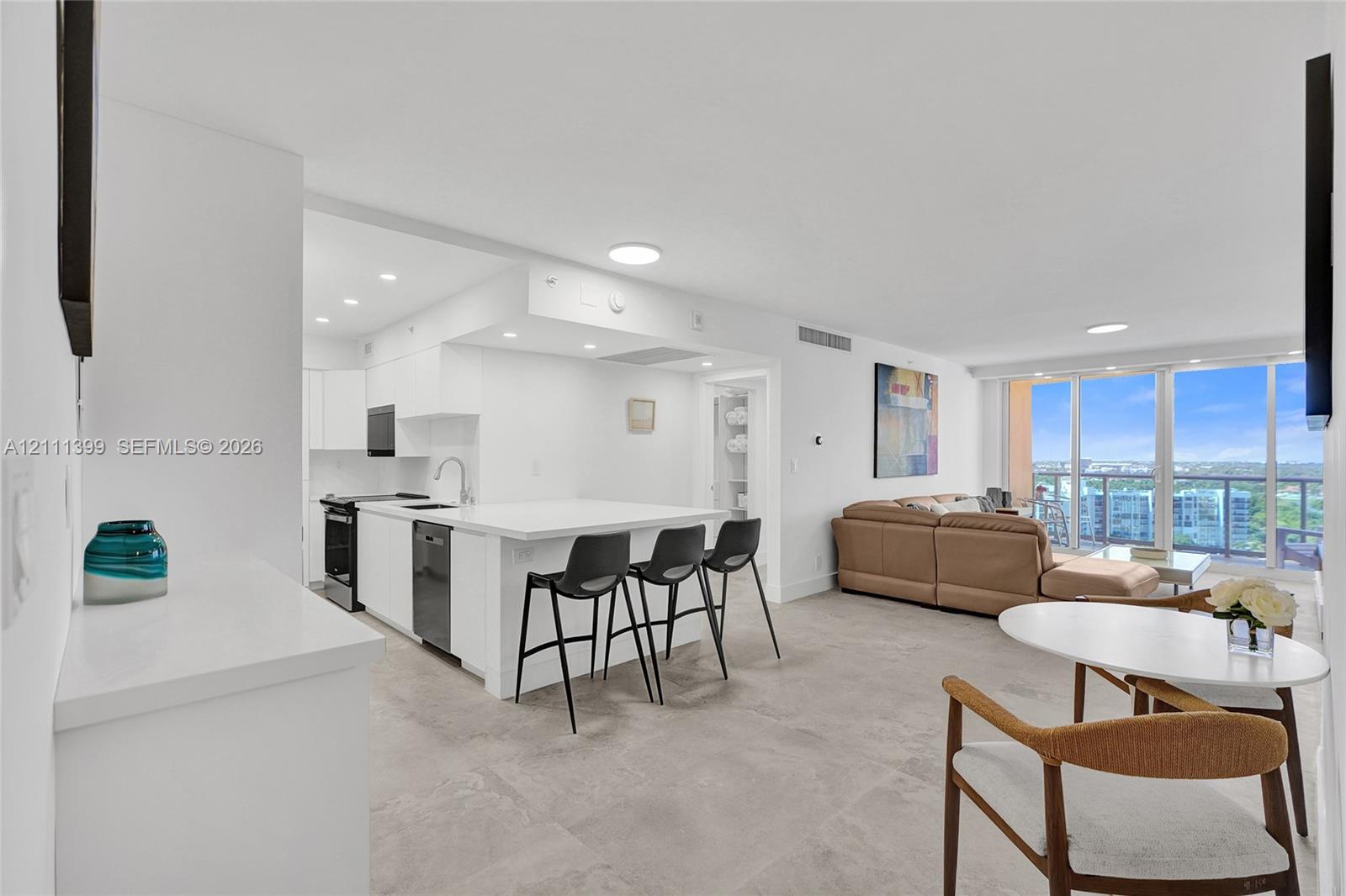 Open-concept kitchen featuring white cabinetry, a stone-finish island with integrated breakfast bar, recessed lighting, and large-format tile flooring, complete with floor-to-ceiling windows