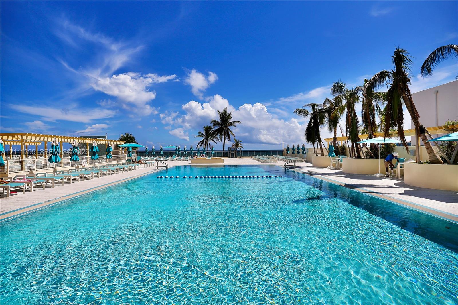 Large rectangular swimming pool featuring a light-colored paved deck, integrated pergola structures, and raised planters with mature palm trees