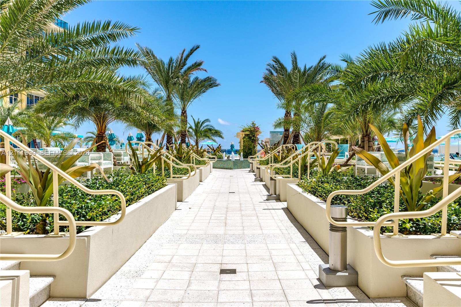 Tiled outdoor promenade featuring custom planters, mature palm trees, integrated metal handrails, and a central water fountain