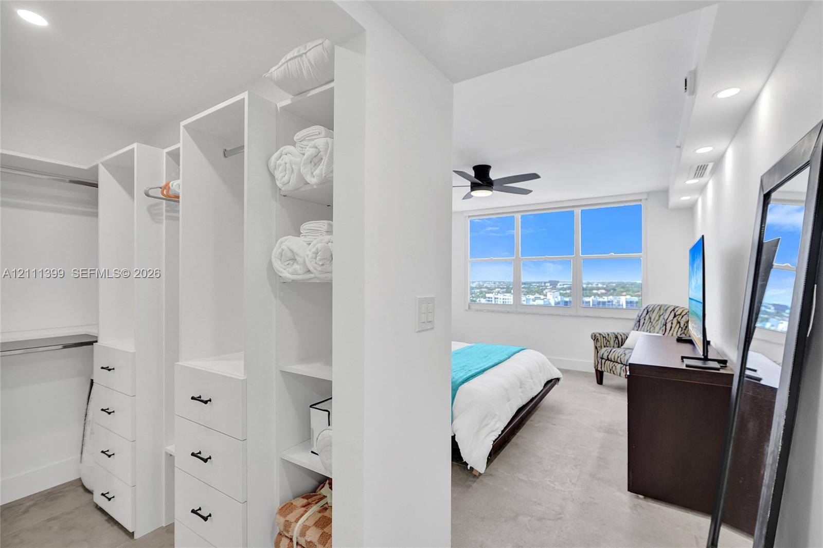 Custom walk-in closet featuring white built-in shelving, hanging rods, and drawers with matte black hardware, recessed ceiling lighting, and large windows with exterior scenery