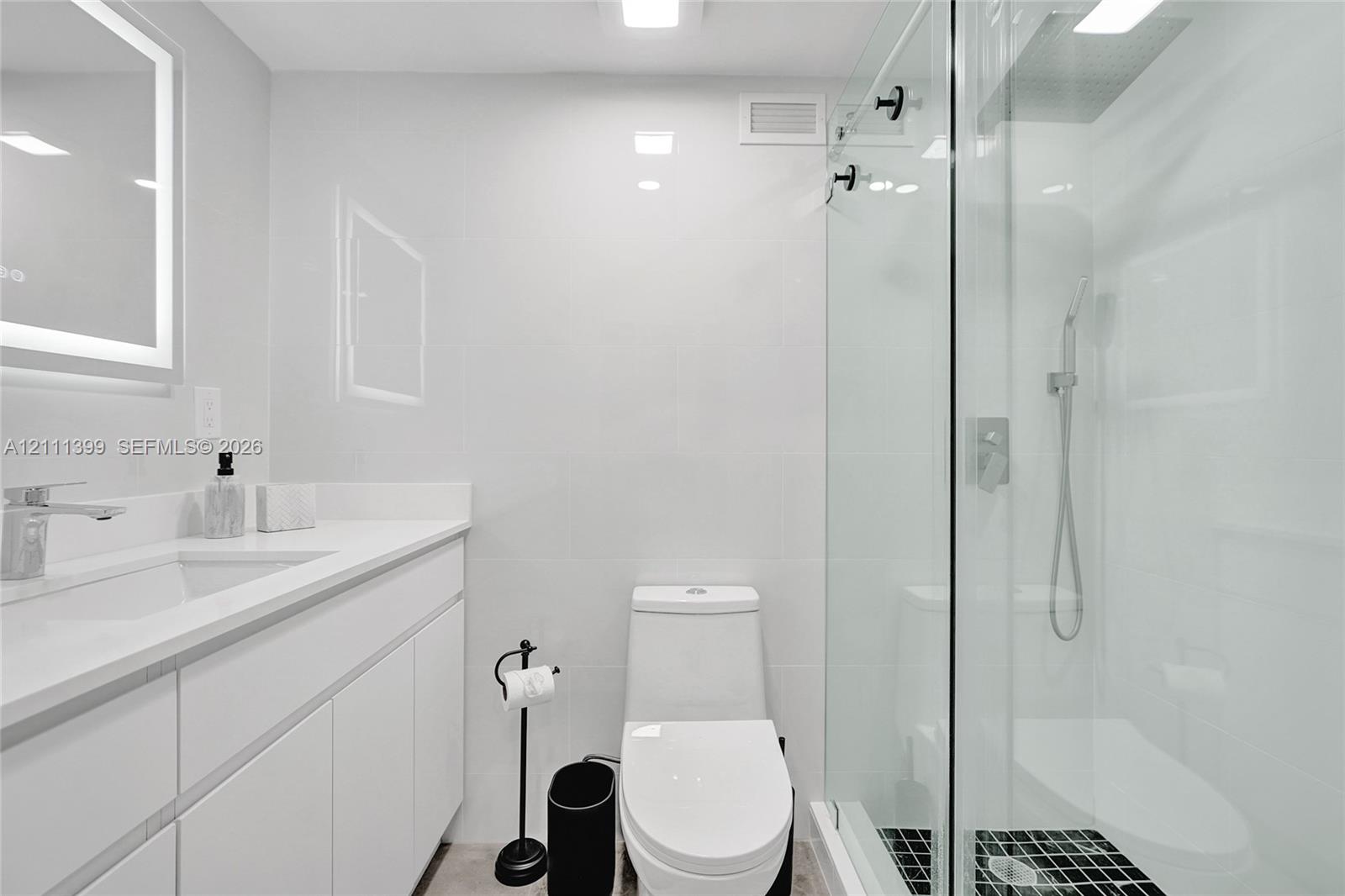 Bathroom featuring a glass-enclosed walk-in shower with a rainfall showerhead, white vanity with integrated basin, polished metal hardware, and LED-backlit mirror