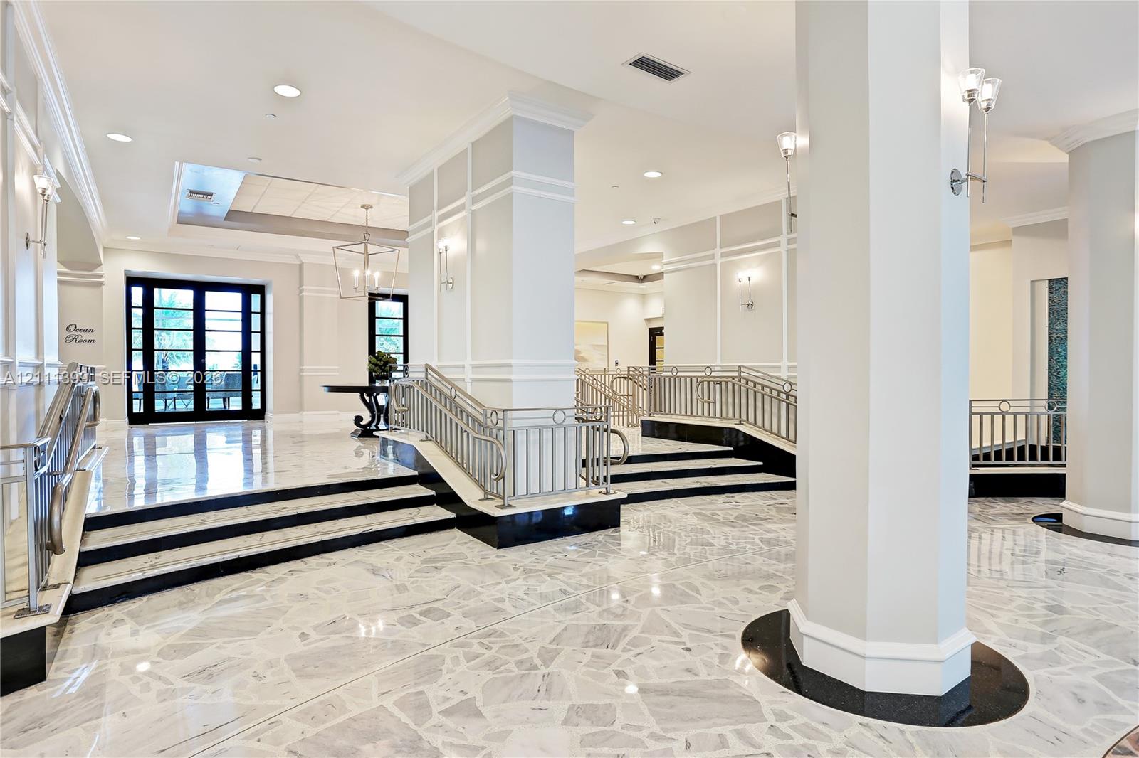 Grand foyer featuring polished stone flooring, custom wall millwork, tiered levels with dark stone accents, metallic railings, and decorative lighting fixtures