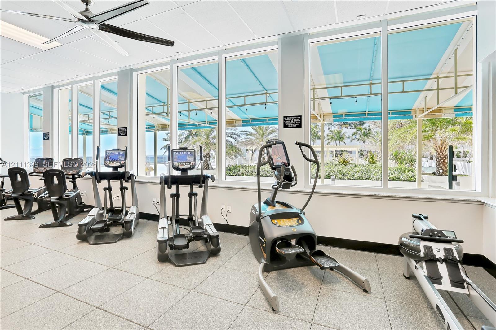 Fitness room featuring floor-to-ceiling windows, large-format tile flooring, a ceiling fan, and external canopy covering