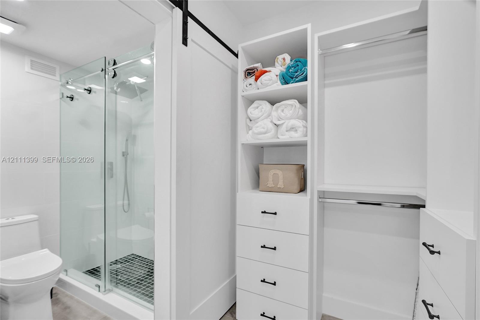 Walk-in shower with a glass enclosure and a black tiled floor, complemented by custom built-in cabinetry featuring white shelving, four storage drawers with matte black hardware, and an integrated hanging rod