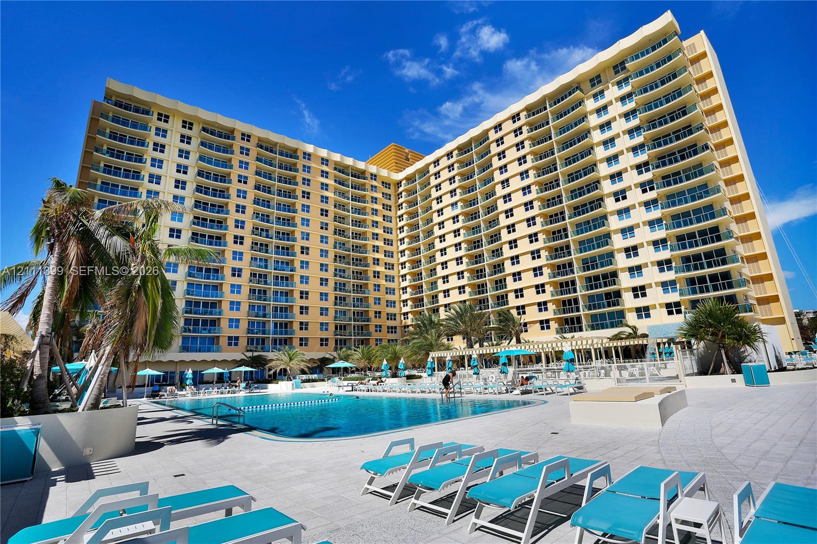 High-rise condominium building featuring tiered private balconies, an expansive outdoor swimming pool, and stone-paved sun deck