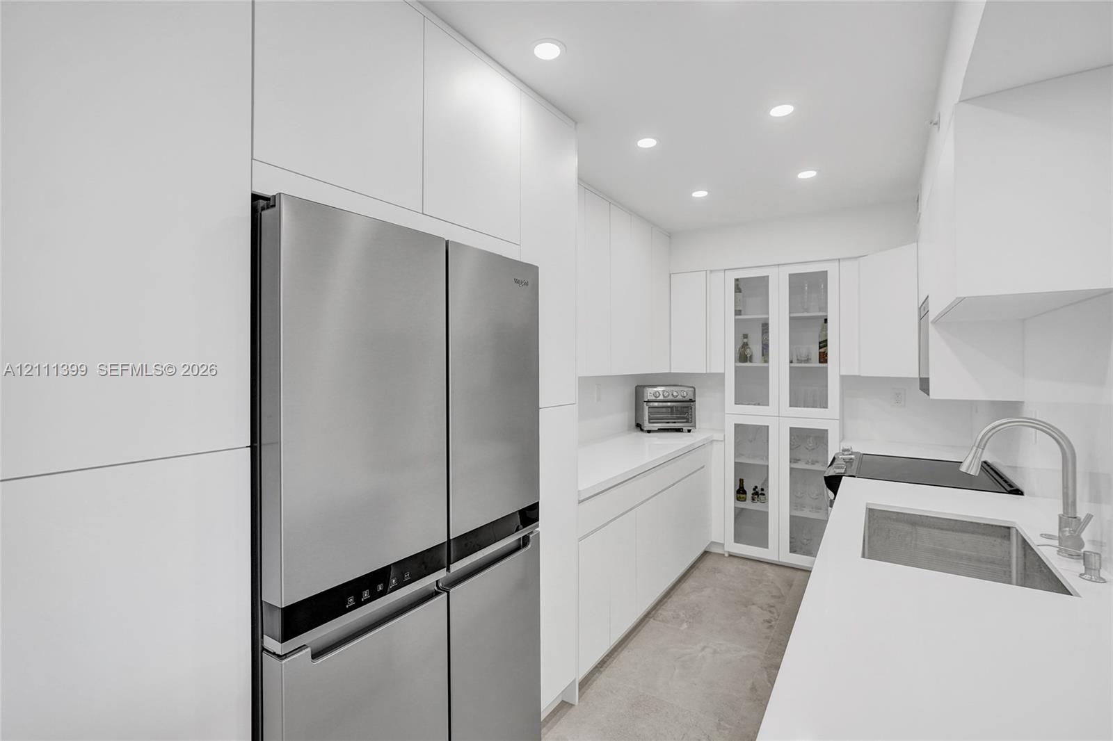 Contemporary kitchen featuring handleless white cabinetry, solid-surface countertops, integrated stainless steel appliances, built-in glass-front display cabinetry, and recessed lighting