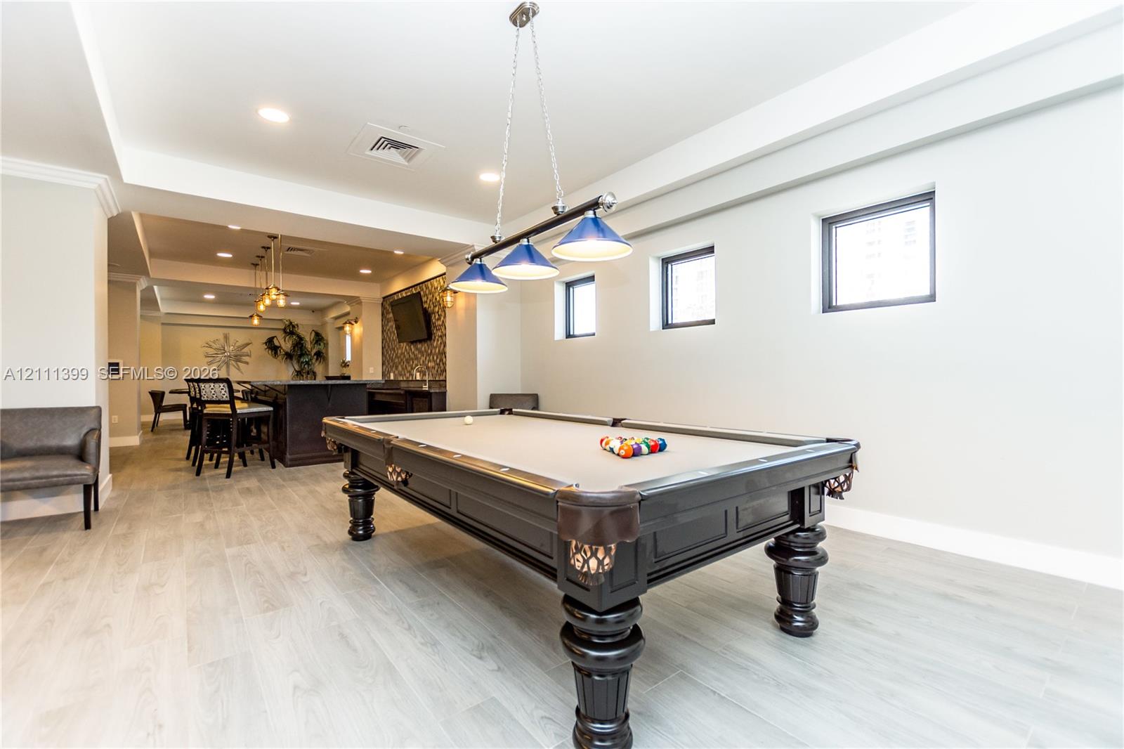 Recessed lighting, wood-finish flooring, three-pendant billiards light fixture, and built-in wet bar featuring dark cabinetry and patterned tile backsplash