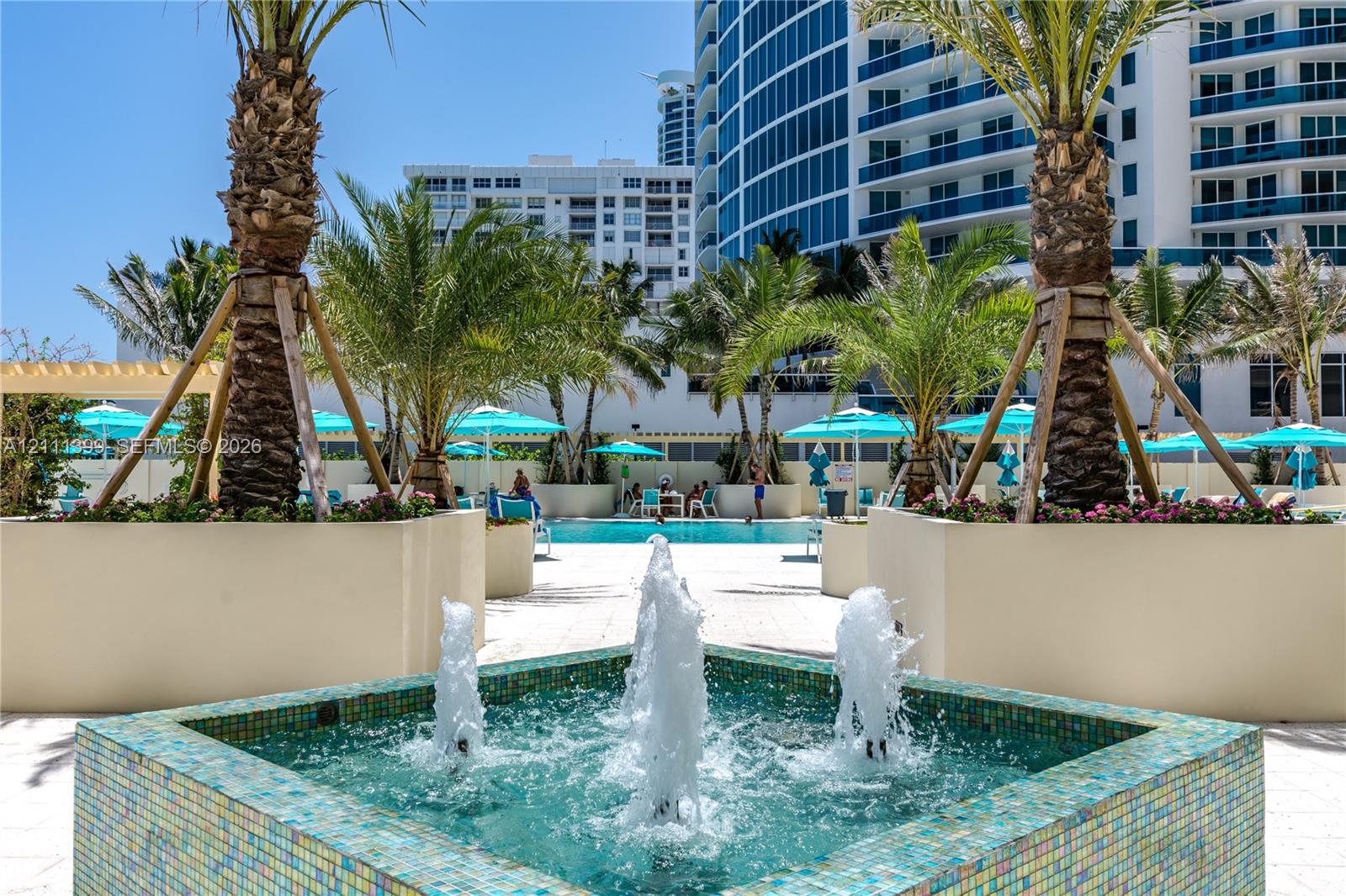 Outdoor swimming pool area featuring a decorative tiled fountain, raised concrete planters, mature palm trees, and stone-finish decking