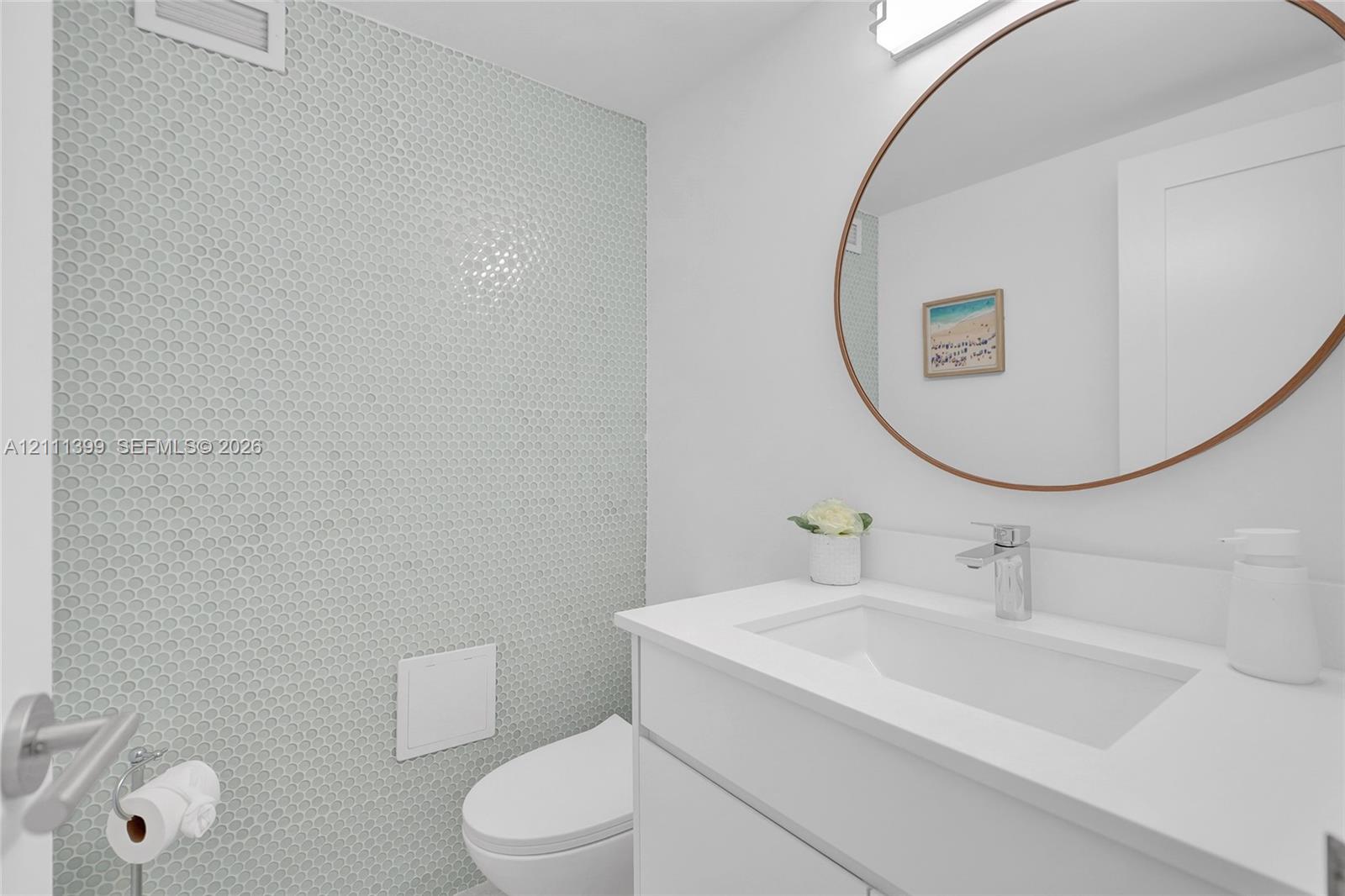 Modern vanity with integrated sink, quartz countertop, and polished chrome faucet, accented by a textured penny tile feature wall and a circular mirror with a bronze frame