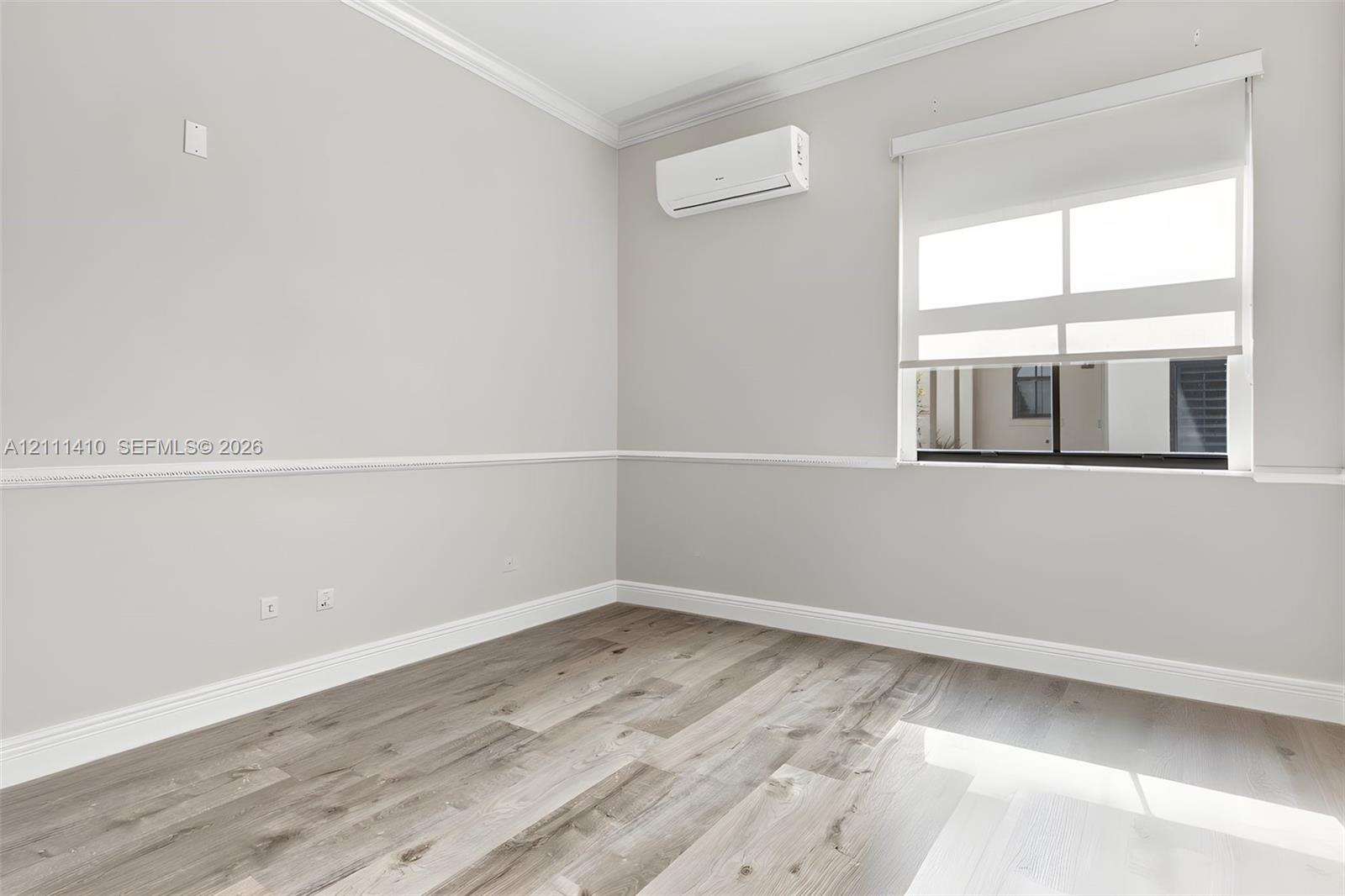 Neutral-toned room featuring wood-finish flooring, chair rail molding, crown molding, and a wall-mounted ductless air conditioning unit, completed by a window with a retractable roller shade