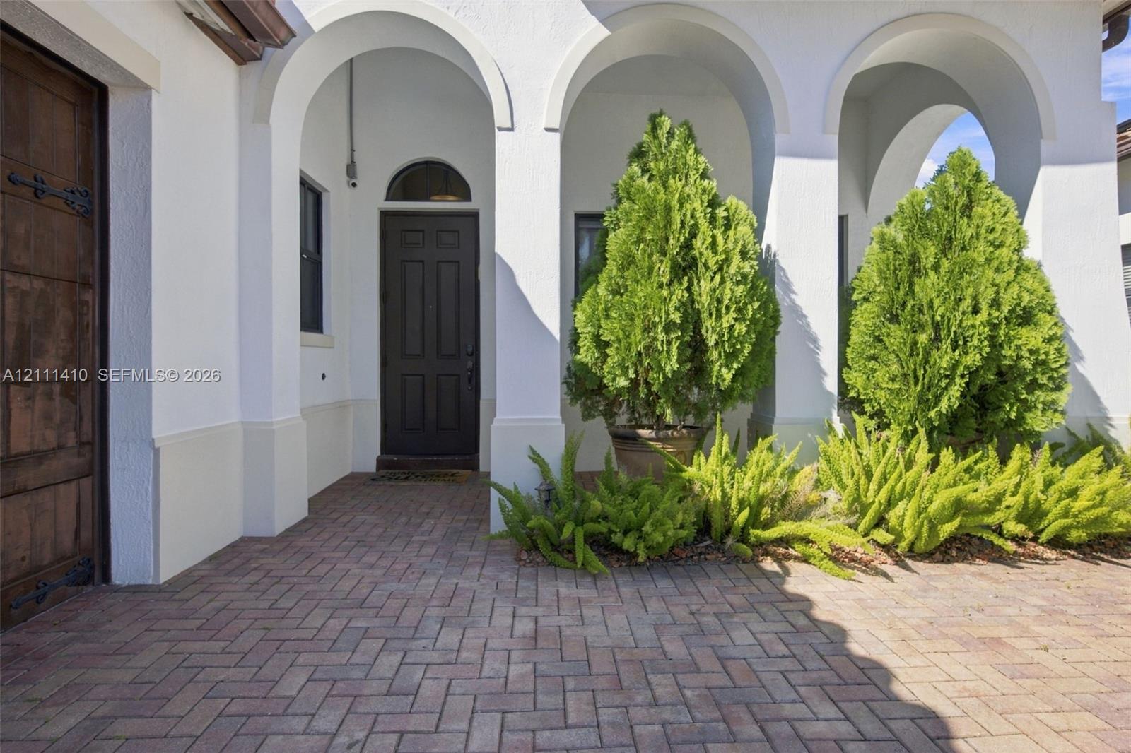 Covered entryway featuring arched architectural detailing, herringbone-patterned brick pavers, a solid wood-finish entry door with metal hardware, and stucco exterior walls Covered entryway featuring arched architectural detailing, herringbone-patterned brick pavers, a solid wood-finish entry door with metal hardware, and stucco exterior walls
