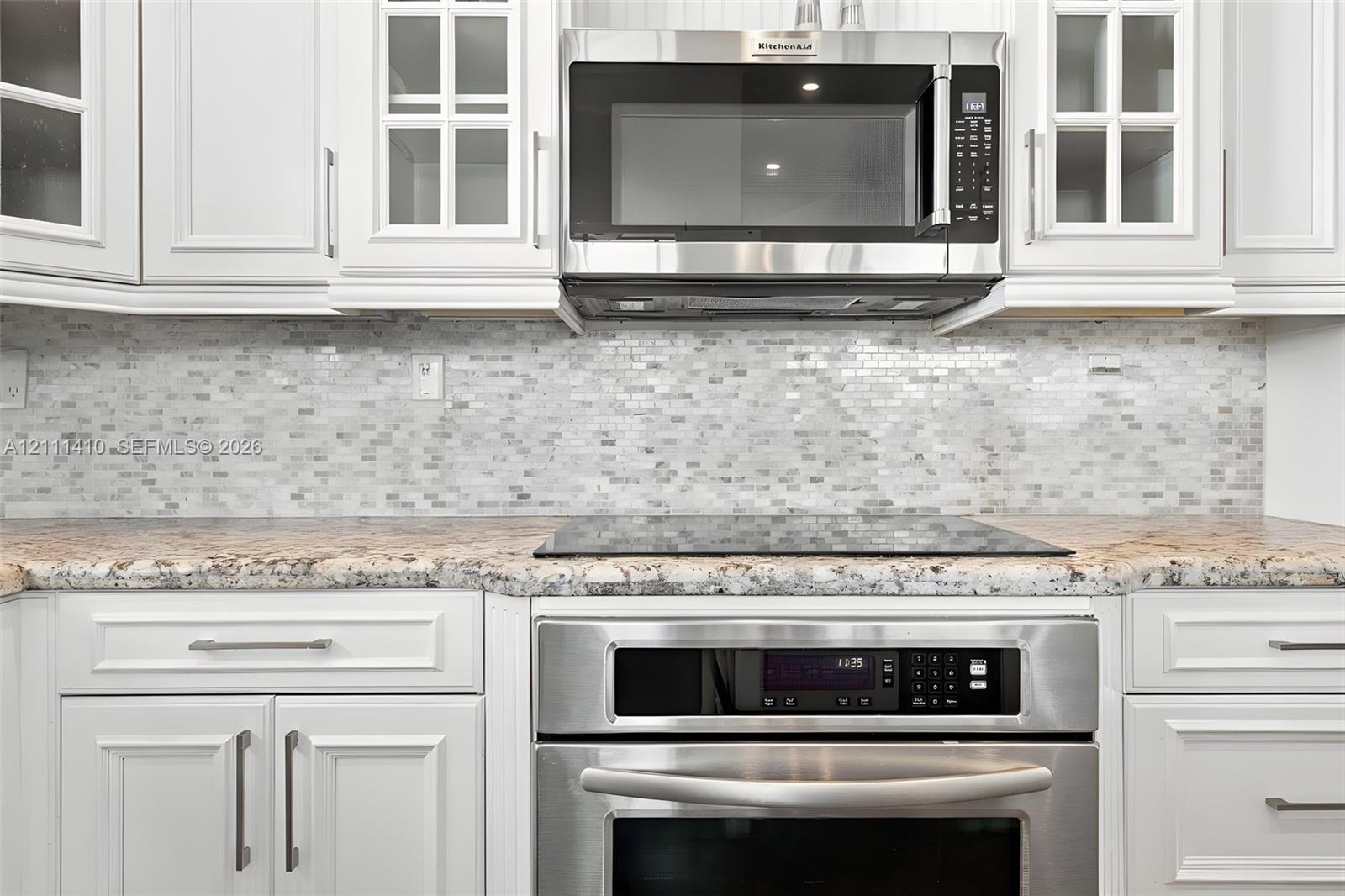 White shaker-style cabinetry with glass-front uppers, stone-finish countertops, mosaic tile backsplash, and stainless steel built-in appliances including a cooktop, wall oven, and microwave