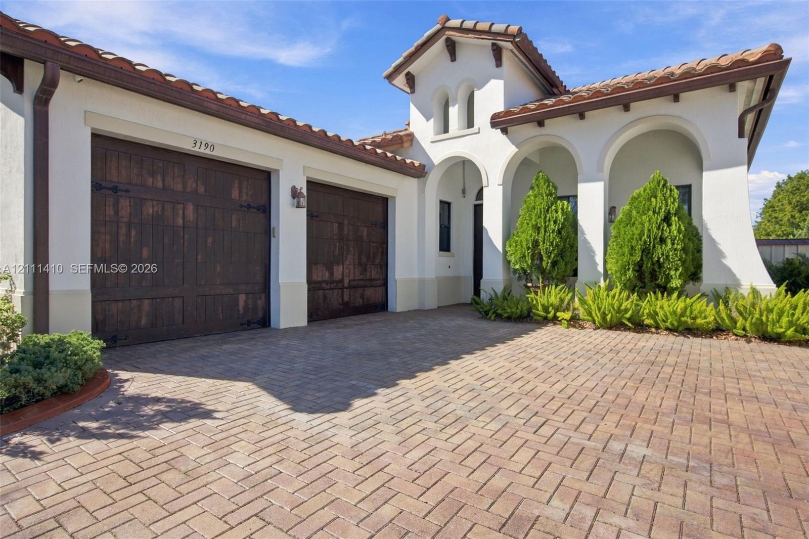 Stucco exterior featuring arched entryways, a terracotta-style tile roof, dual wood-finish garage doors, and a herringbone-patterned paver driveway Stucco exterior featuring arched entryways, a terracotta-style tile roof, dual wood-finish garage doors, and a herringbone-patterned paver driveway