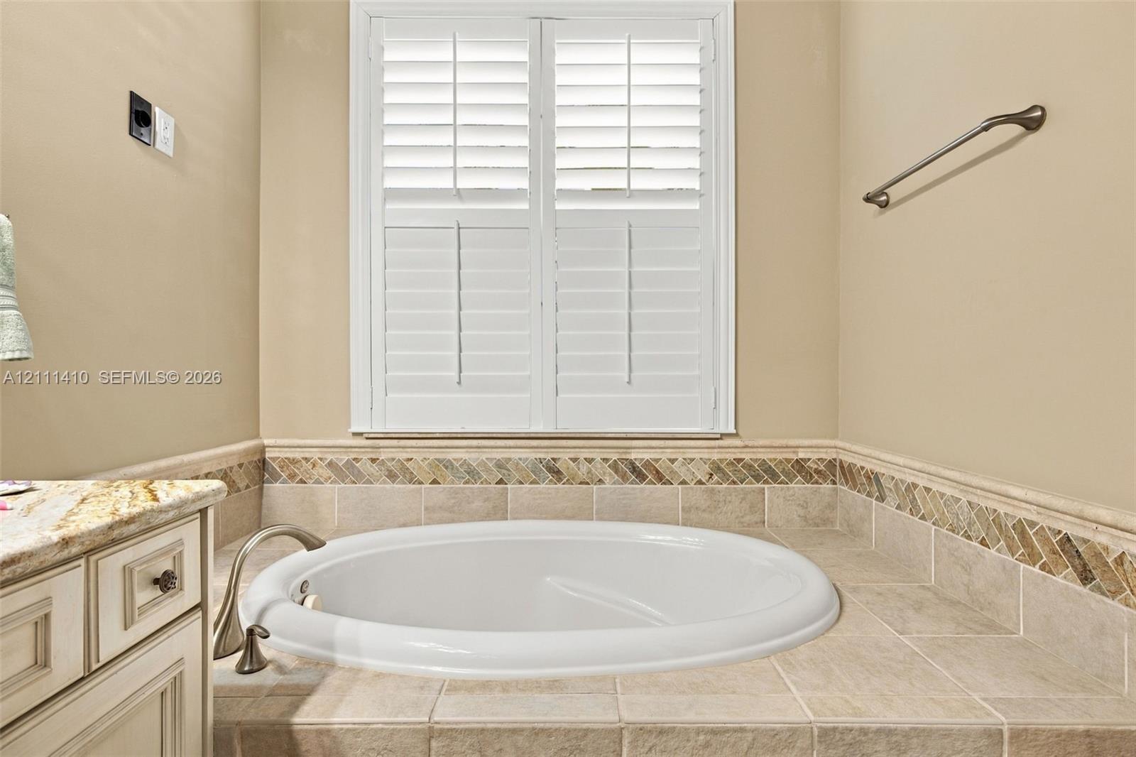 Built-in soaking tub with a tile surround, herringbone-patterned stone backsplash, plantation shutters, and a vanity featuring a stone countertop