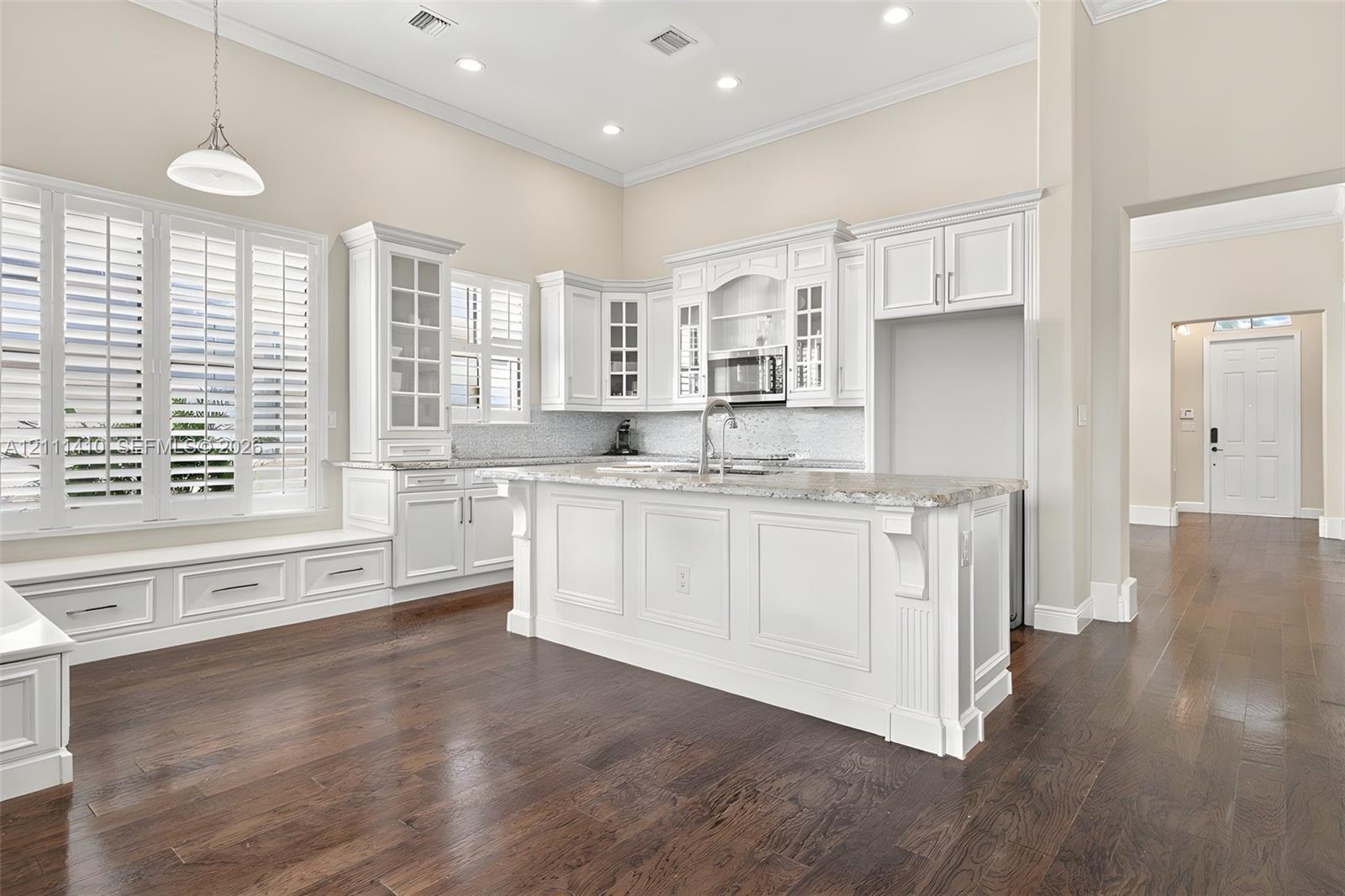 Kitchen featuring white cabinetry with glass-front uppers, a central island with stone-finish countertops, recessed lighting, plantation shutters, and wood-finish flooring Kitchen featuring white cabinetry with glass-front uppers, a central island with stone-finish countertops, recessed lighting, plantation shutters, and wood-finish flooring