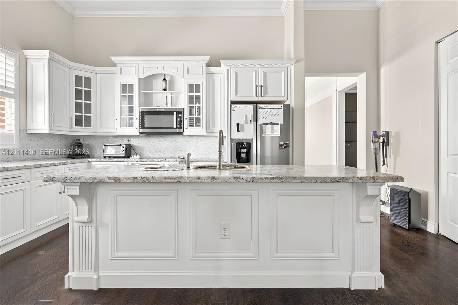 Kitchen featuring white cabinetry with crown molding, stone countertops, a large center island with corbel detailing, stainless steel appliances, and dark wood-finish flooring Kitchen featuring white cabinetry with crown molding, stone countertops, a large center island with corbel detailing, stainless steel appliances, and dark wood-finish flooring
