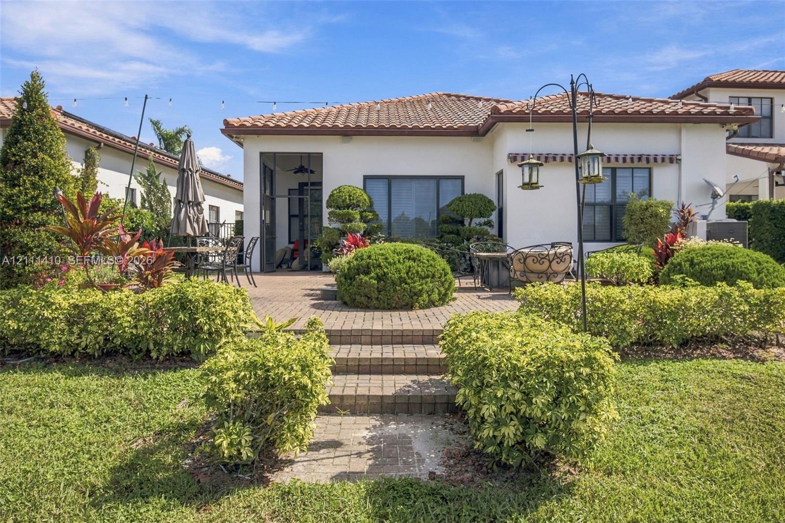 Rear exterior featuring a tile roof, paved terrace, multi-level stairs, manicured landscaping, and a screened enclosure