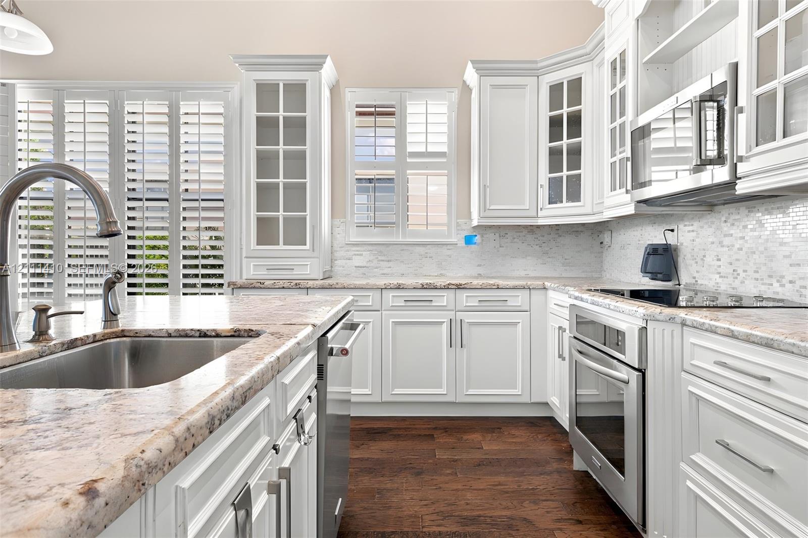 Kitchen featuring custom white cabinetry with glass-front uppers, stone countertops, stainless steel appliances, a mosaic tile backsplash, and wood-finish flooring Kitchen featuring custom white cabinetry with glass-front uppers, stone countertops, stainless steel appliances, a mosaic tile backsplash, and wood-finish flooring