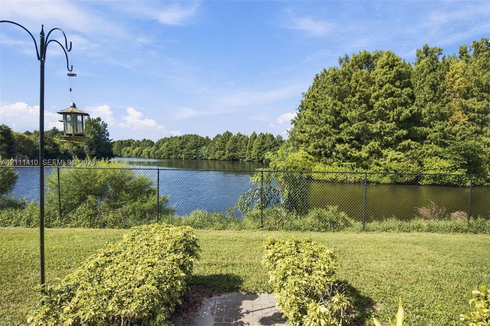 Backyard featuring a manicured lawn, established shrubbery, a black metal chain-link fence, and direct water frontage with dense natural perimeter trees