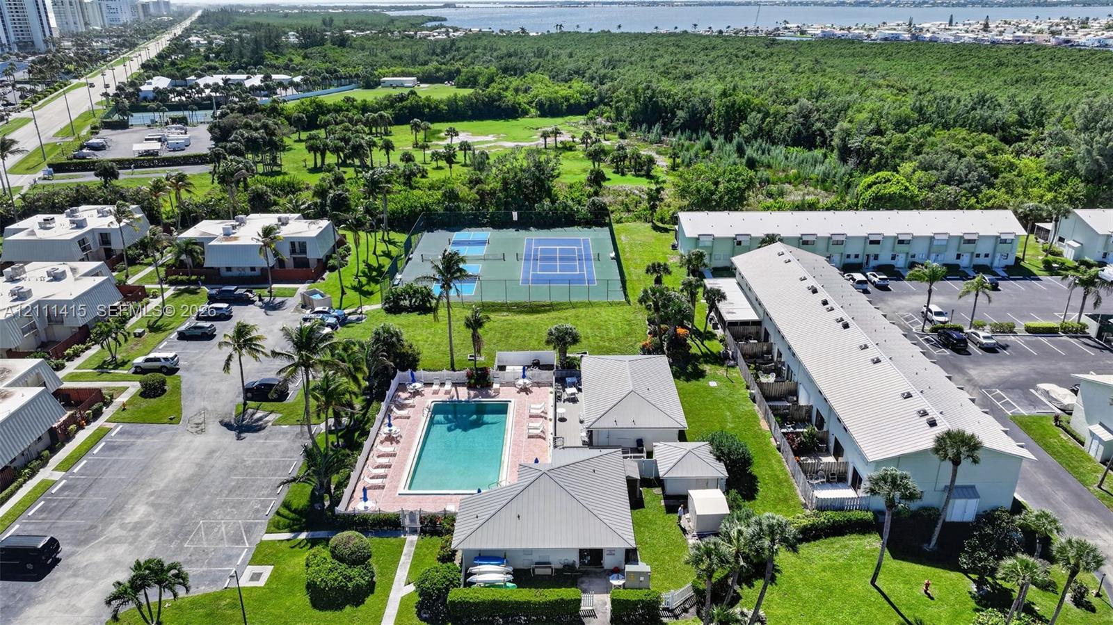 Aerial perspective of multi-family complex featuring a swimming pool with paved deck, two fenced tennis courts, expansive green space, and mature palm trees