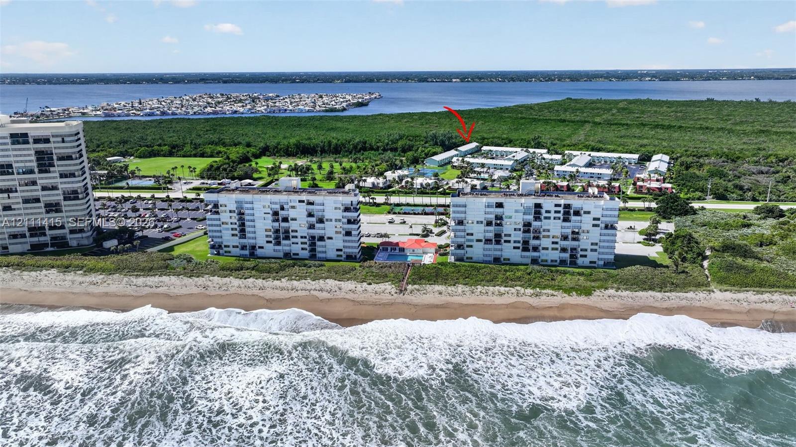 Aerial perspective featuring multi-story residential buildings, direct oceanfront placement, paved parking facilities, and proximity to coastal vegetation and waterway