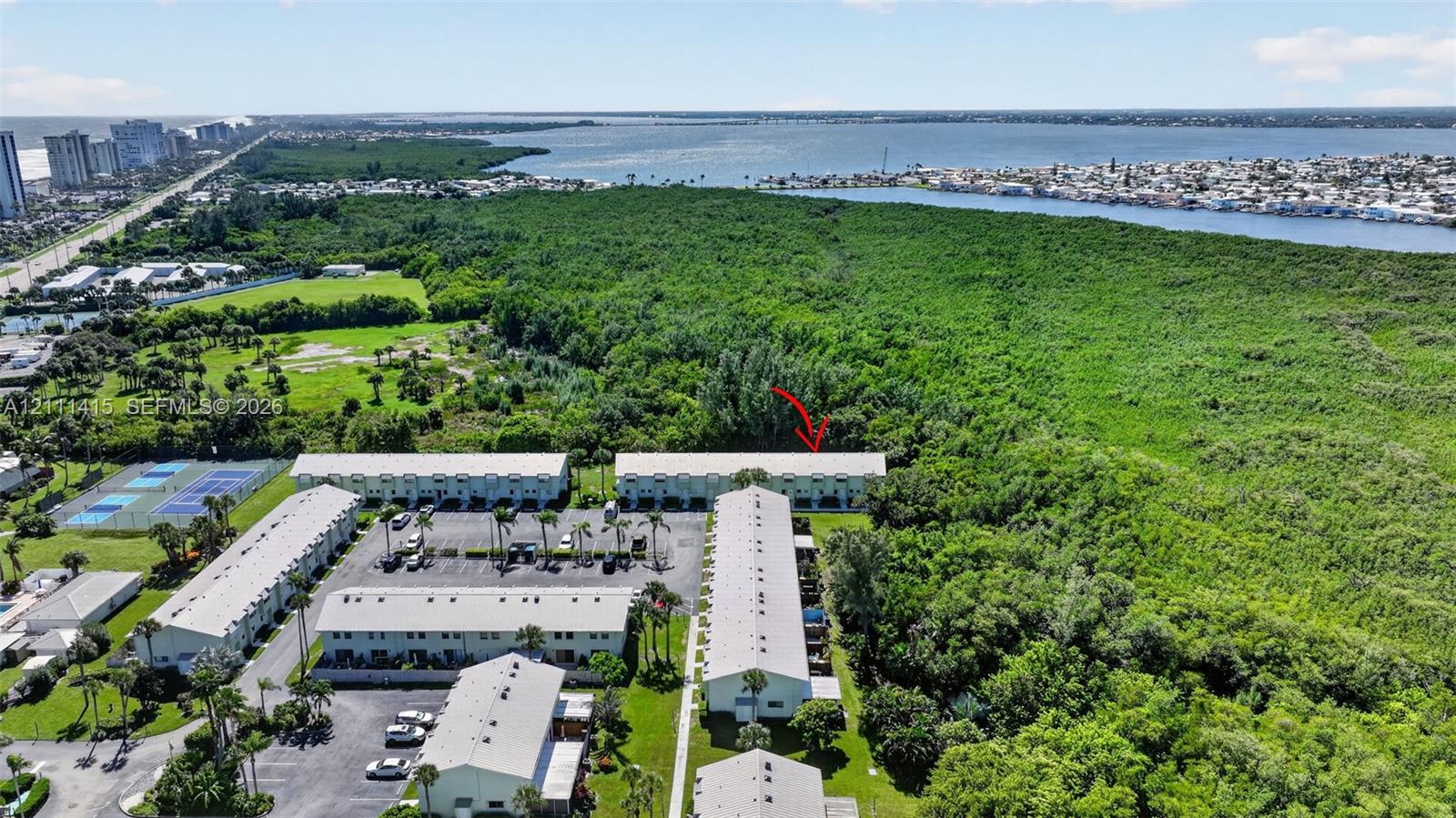 Aerial perspective of a multi-unit complex featuring surface parking, on-site tennis courts, established landscaping, and proximity to coastal waterways