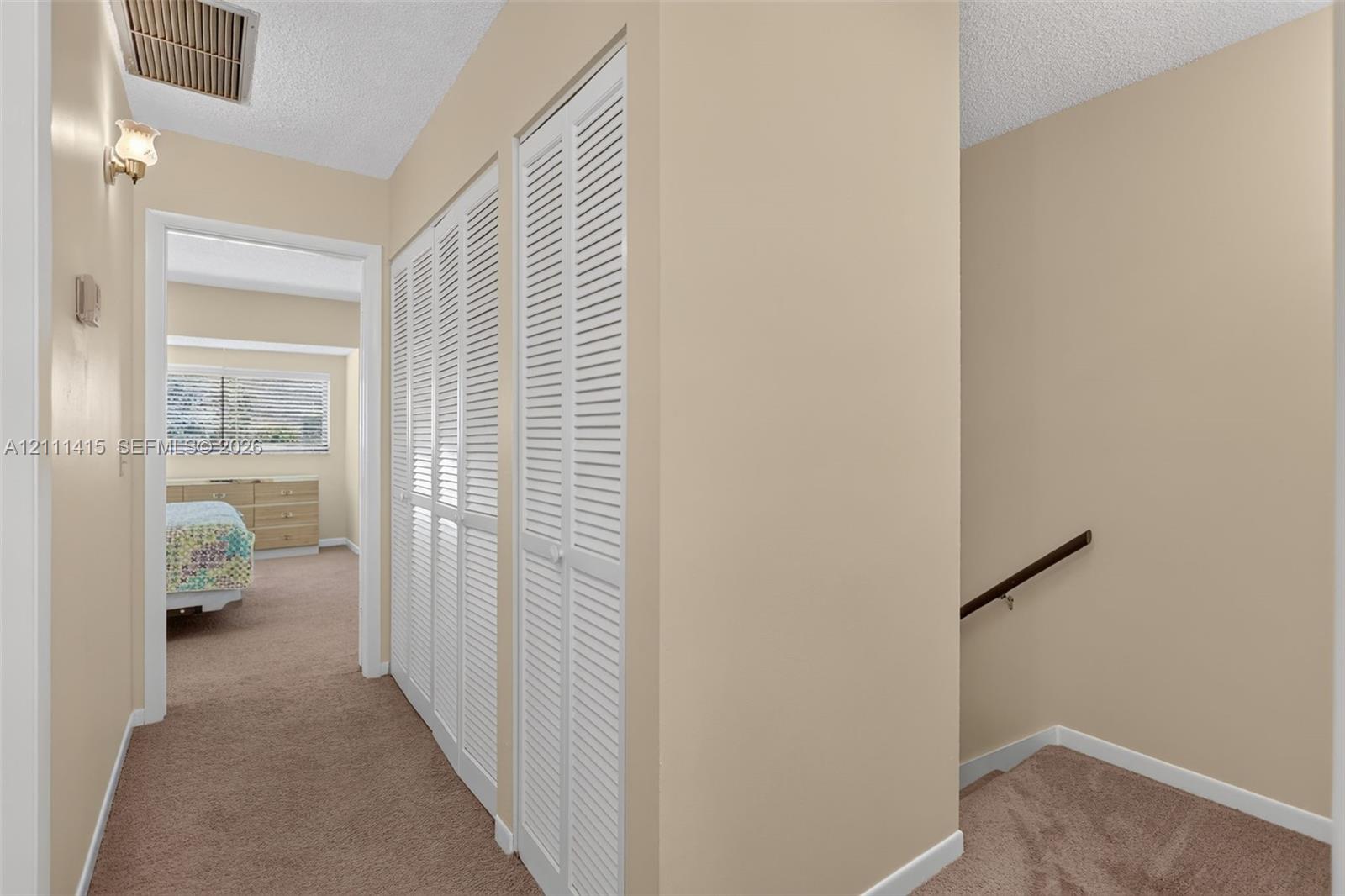 Hallway featuring dual white louvered closet doors, carpeted flooring, and a wall-mounted handrail along the stairwell