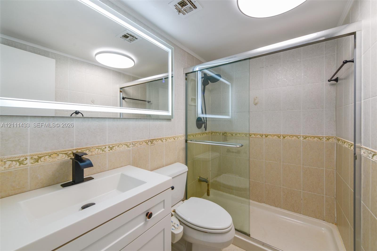 Bathroom featuring a white vanity with integrated basin, matte black faucet, illuminated LED mirror, and a walk-in shower enclosure with sliding glass doors and tiled surround