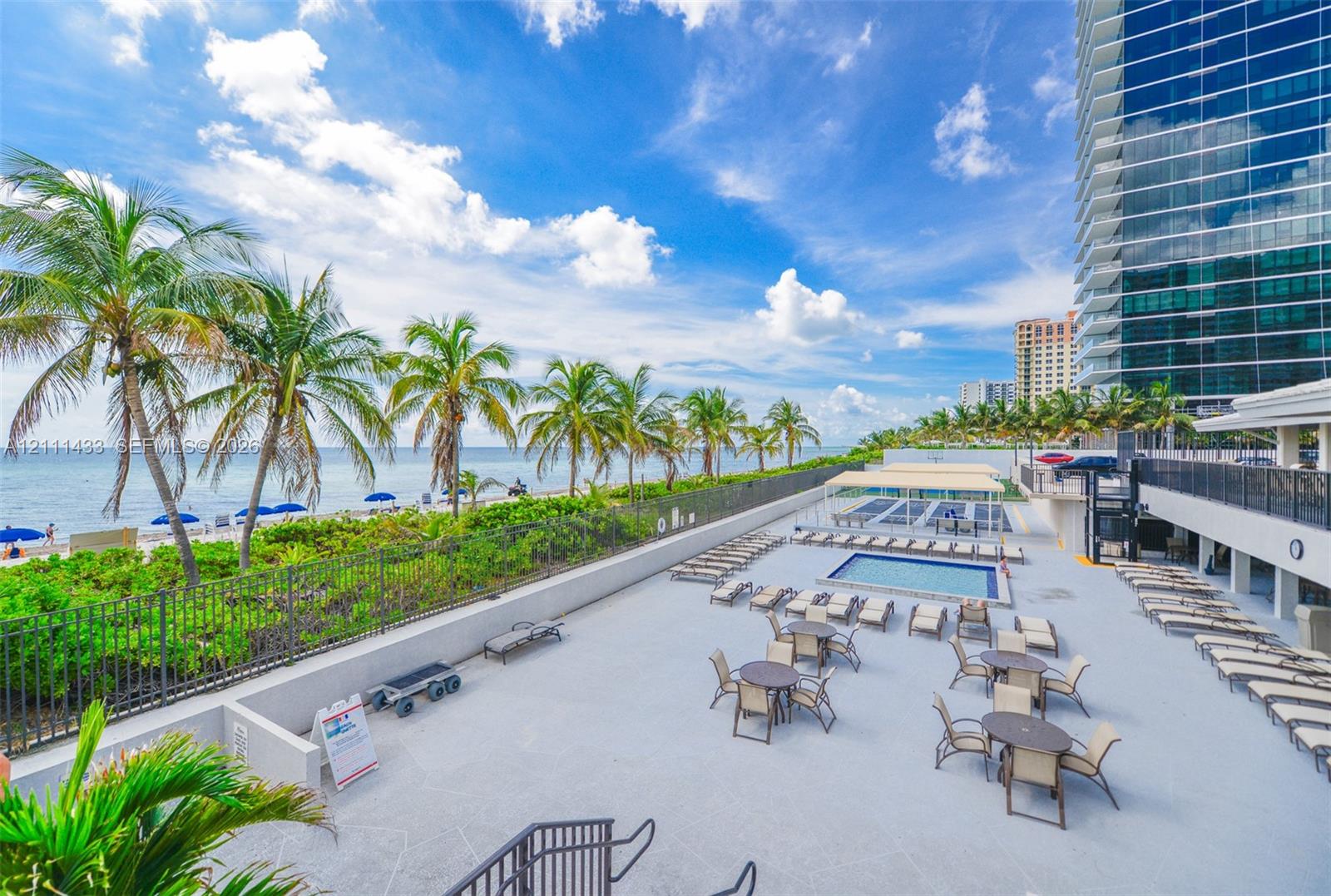 Expansive paved terrace featuring a swimming pool, shaded seating areas, metal perimeter fencing, and mature palm trees