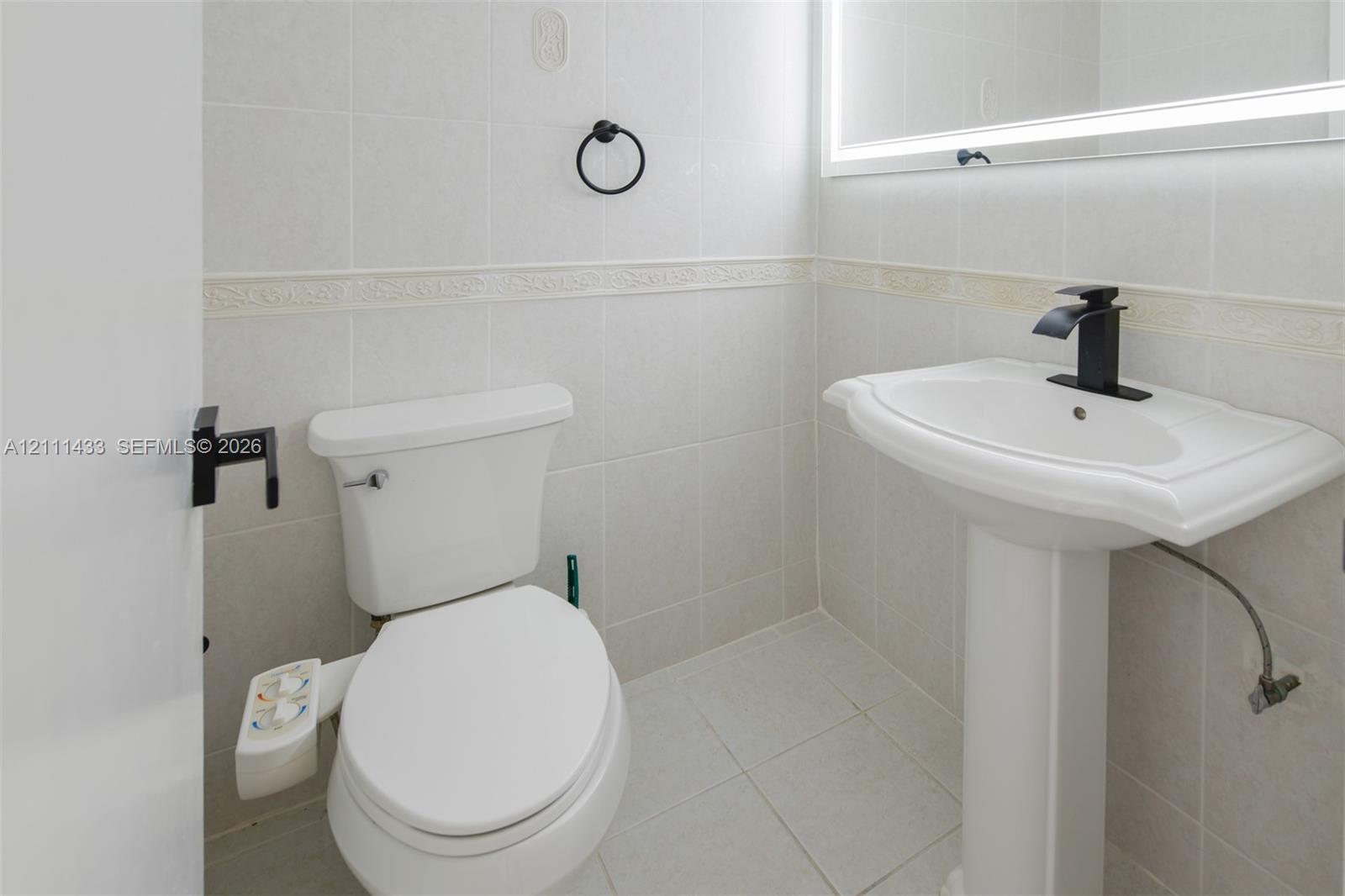 Pedestal sink with a matte black faucet, wall-mounted illuminated mirror, floor-to-ceiling tiled walls with decorative trim, and matte black hardware