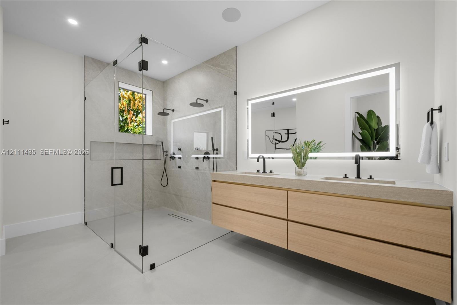 Floating dual-sink vanity with light wood cabinetry and solid-surface countertop, frameless glass walk-in shower with stone tile surround, integrated recessed shower niche, and rectangular wall-mounted backlit mirror