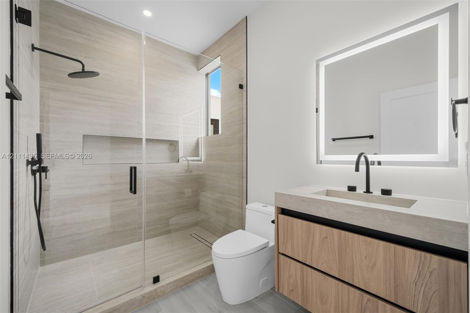 Bathroom featuring a floating wood-finish vanity, integrated stone-composite countertop, illuminated mirror, walk-in shower with floor-to-ceiling tile, and matte black fixtures