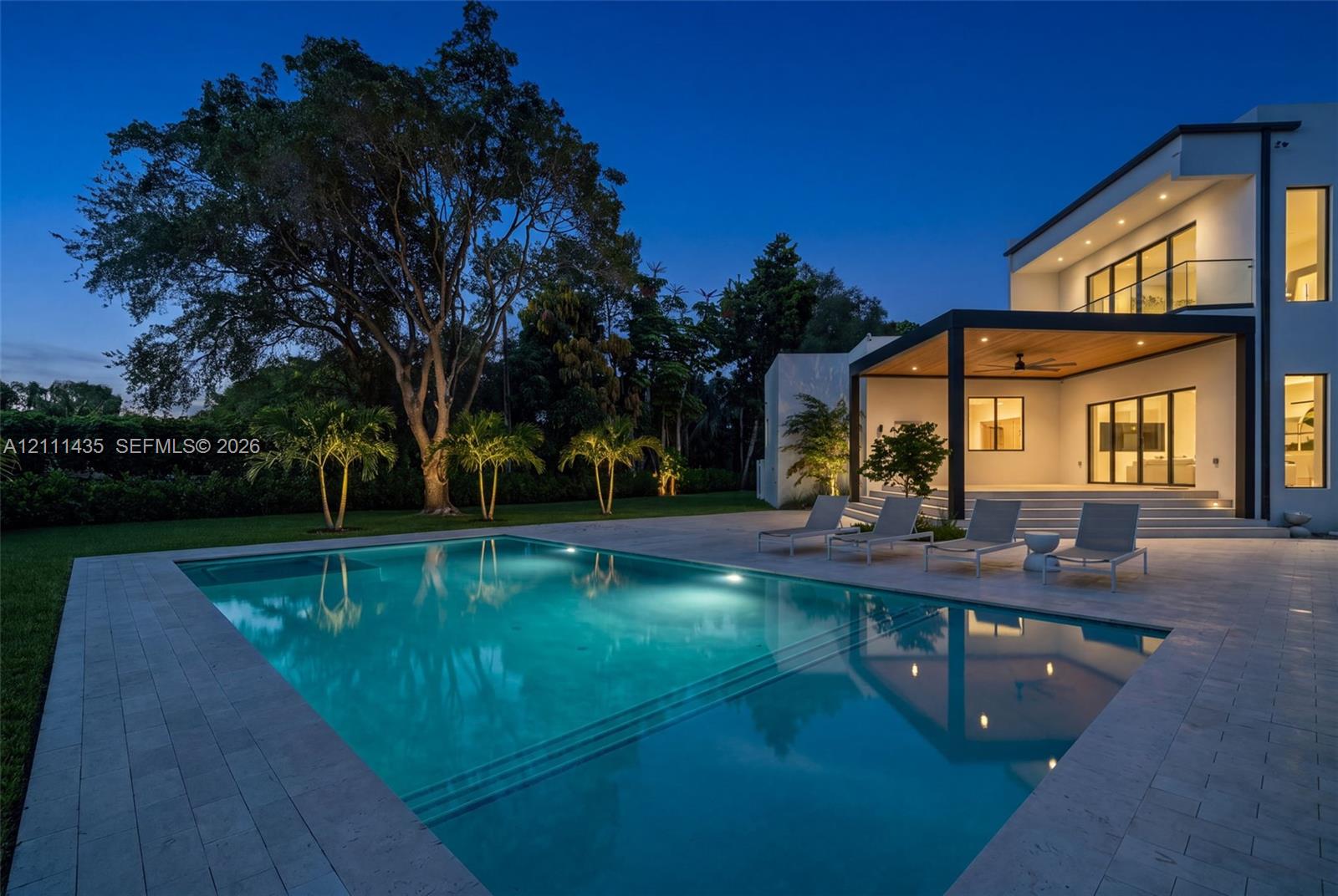 Modern architectural exterior featuring a swimming pool with integrated stairs, expansive stone paver pool deck, covered patio with wood-paneled ceiling, and second-story balcony with glass railing