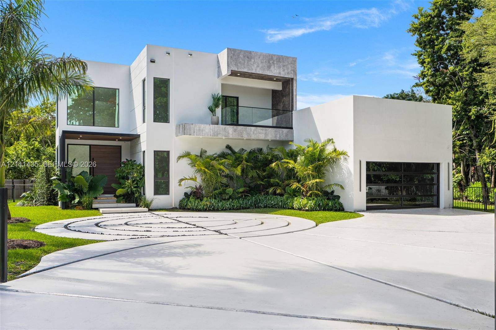 Modern architectural exterior featuring a smooth stucco facade, upper-level terrace with glass railing, decorative circular paver hardscaping, and a multi-panel glass garage door Modern architectural exterior featuring a smooth stucco facade, upper-level terrace with glass railing, decorative circular paver hardscaping, and a multi-panel glass garage door