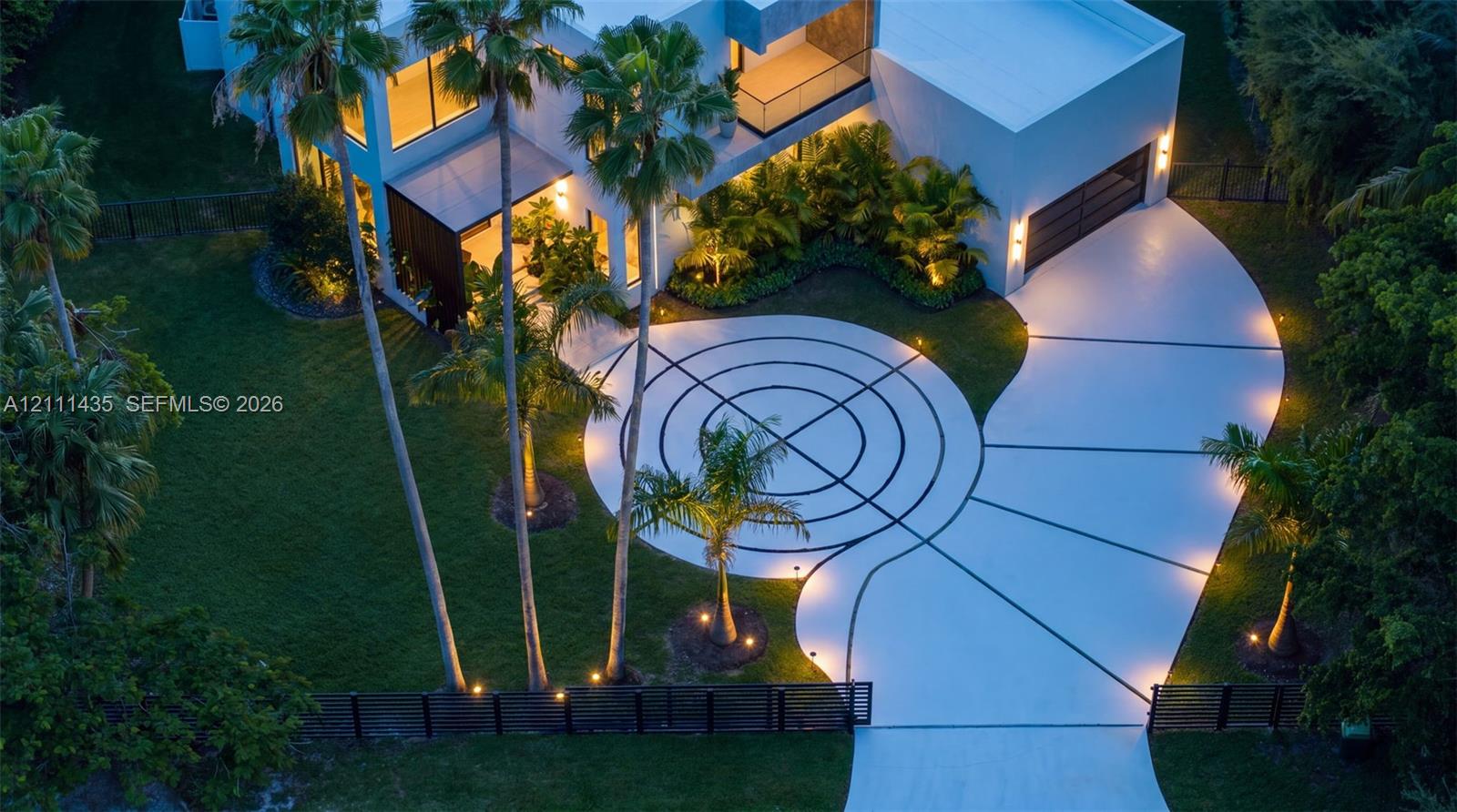 Contemporary architectural exterior featuring a geometric patterned driveway, expansive manicured lawn, multi-level structure with integrated lighting, and mature palm landscaping