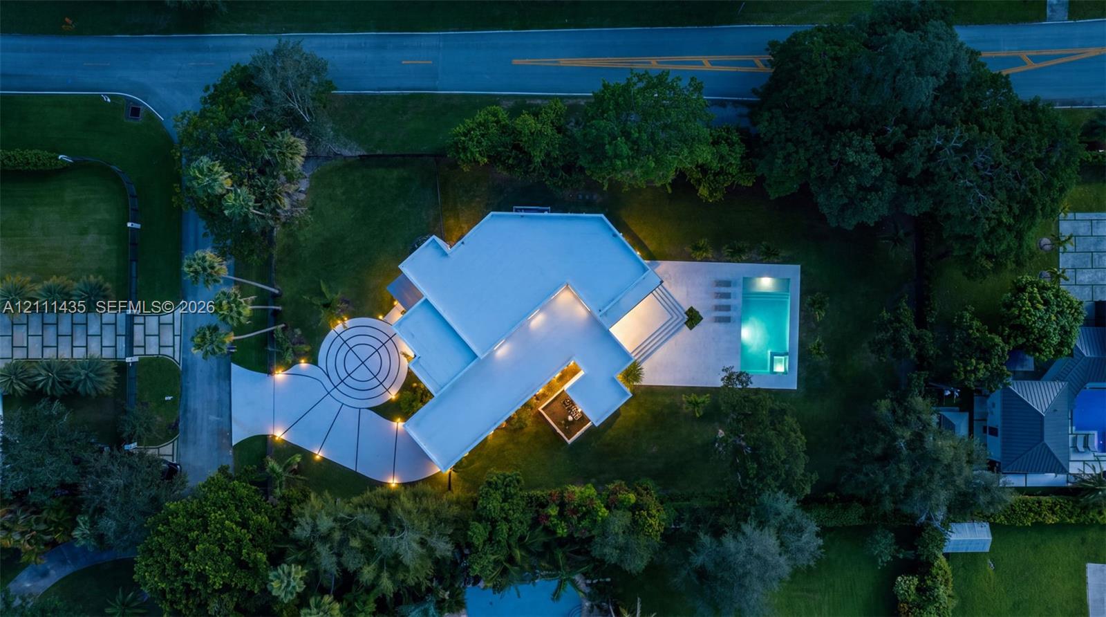 Aerial view featuring a circular concrete driveway, expansive lawn, mature tree canopy, and a rectangular swimming pool with paved terrace